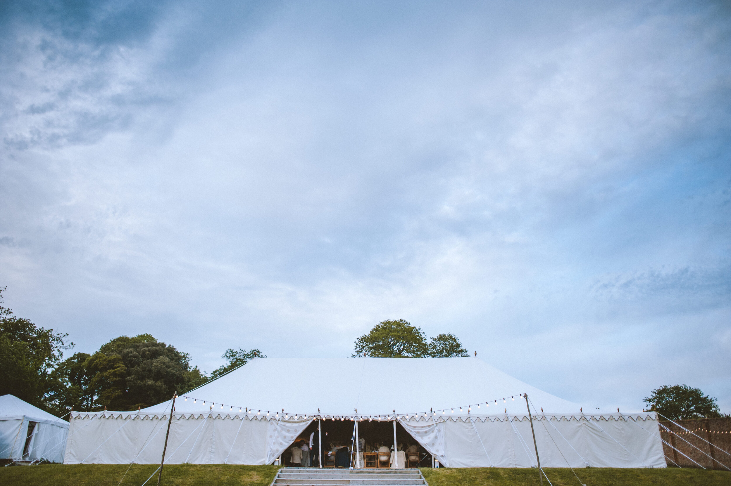 Large white event tent on a grassy area with string lights, set up outdoors under a cloudy sky, with trees in the background.