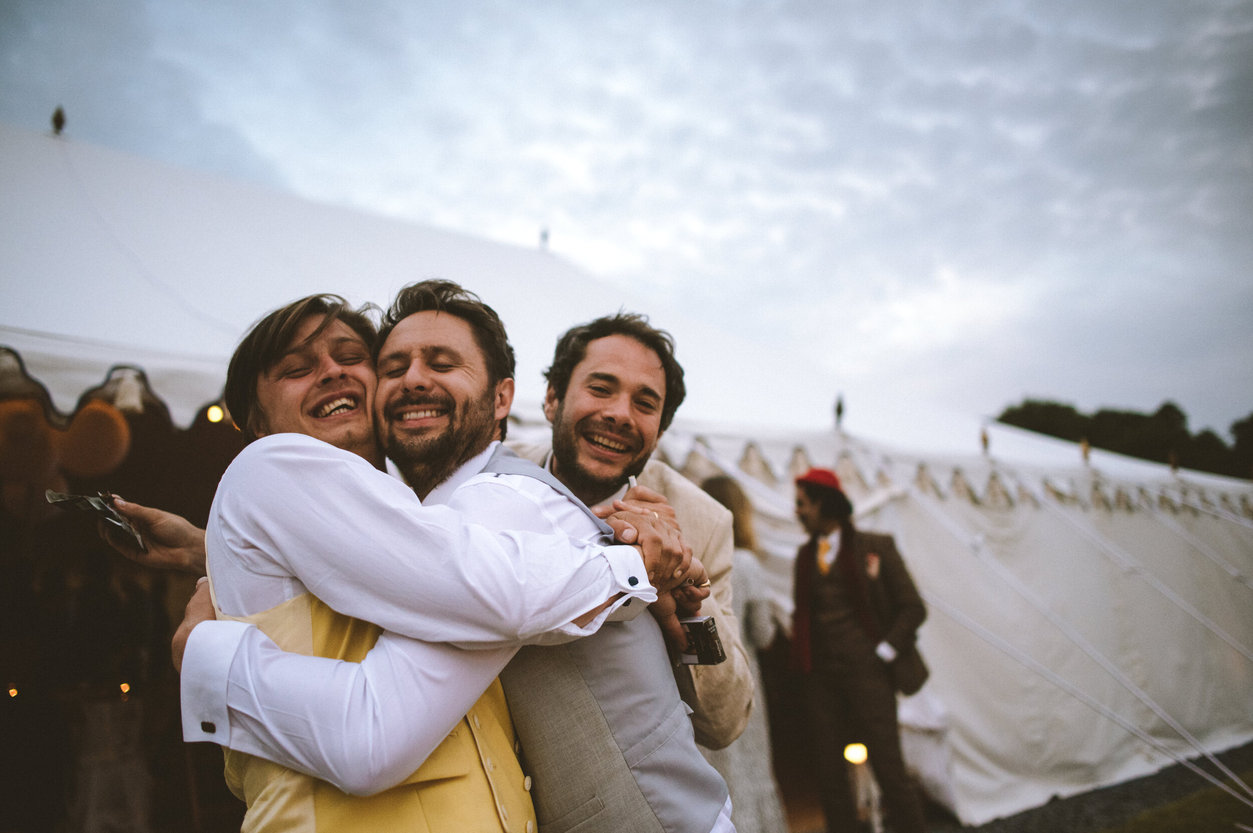 Three men hugging and smiling at an outdoor event, with a large white tent and another person in the background.