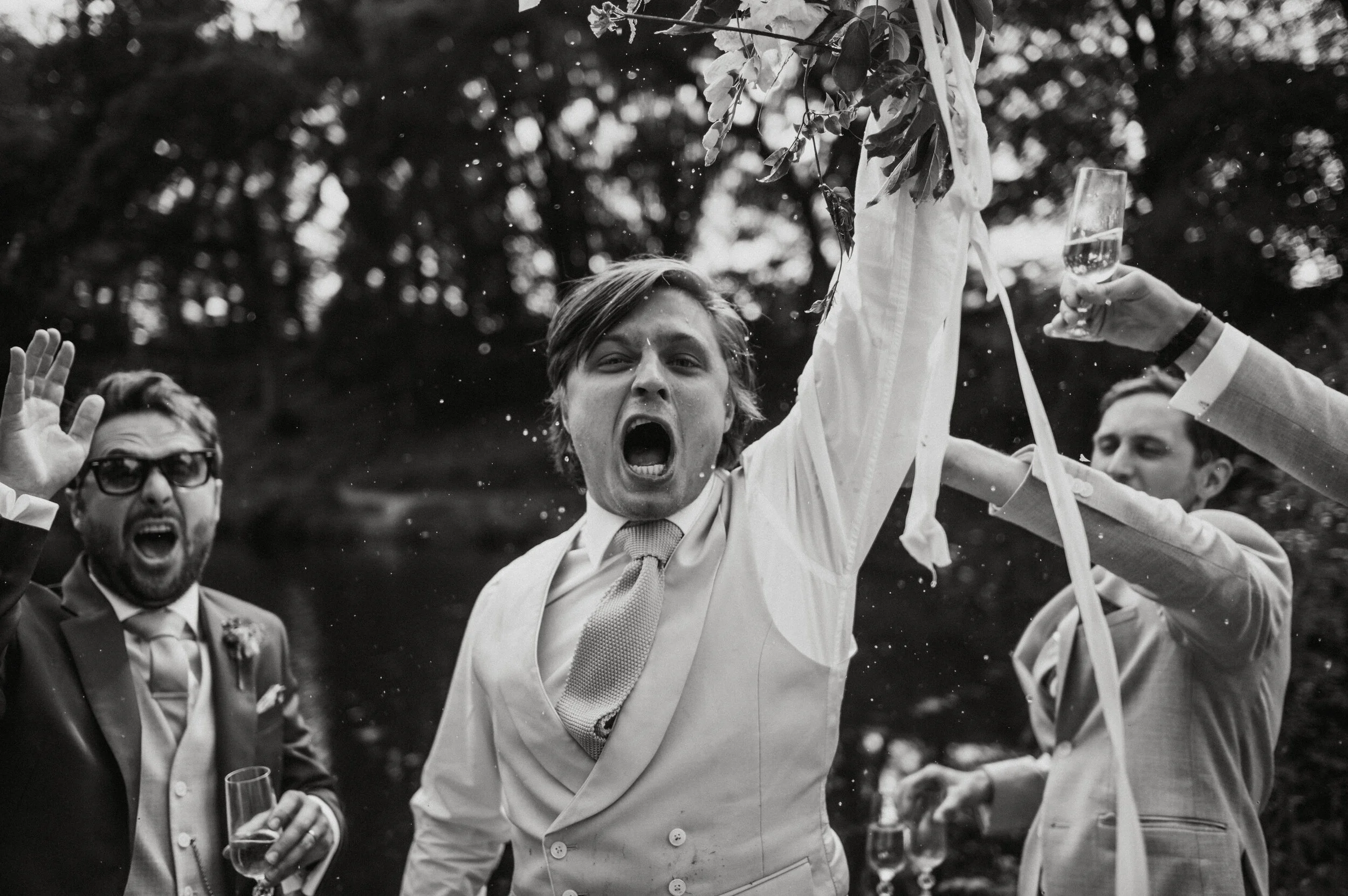 A group of men celebrating outdoors, dressed in suits, with one man in the center shouting joyfully, holding a ribbon. Others are holding drinks, with trees in the background.