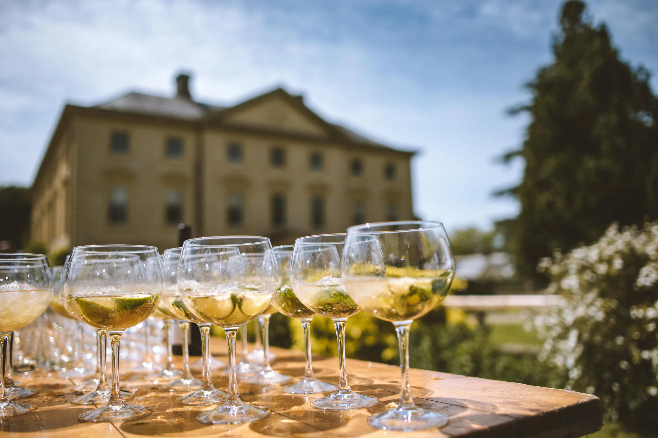 Pencarrow House wedding photography in Cornwall - Multiple glasses of alcohol with lime slices on a wooden table outdoors, with a historic building and trees in the background during daytime.