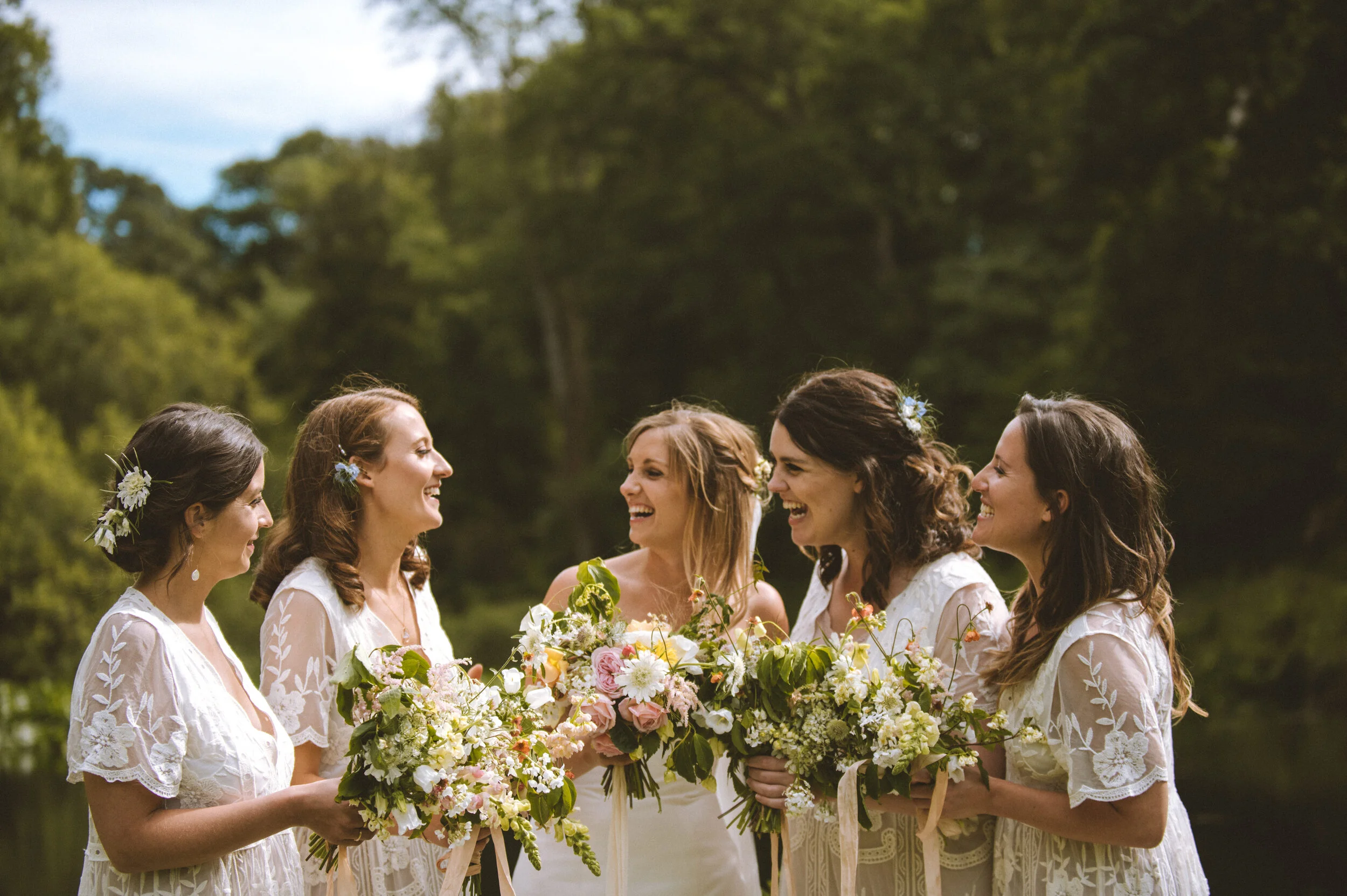 Six women in white dresses holding bouquets of flowers, standing outdoors near a river with trees in the background, smiling and laughing together.