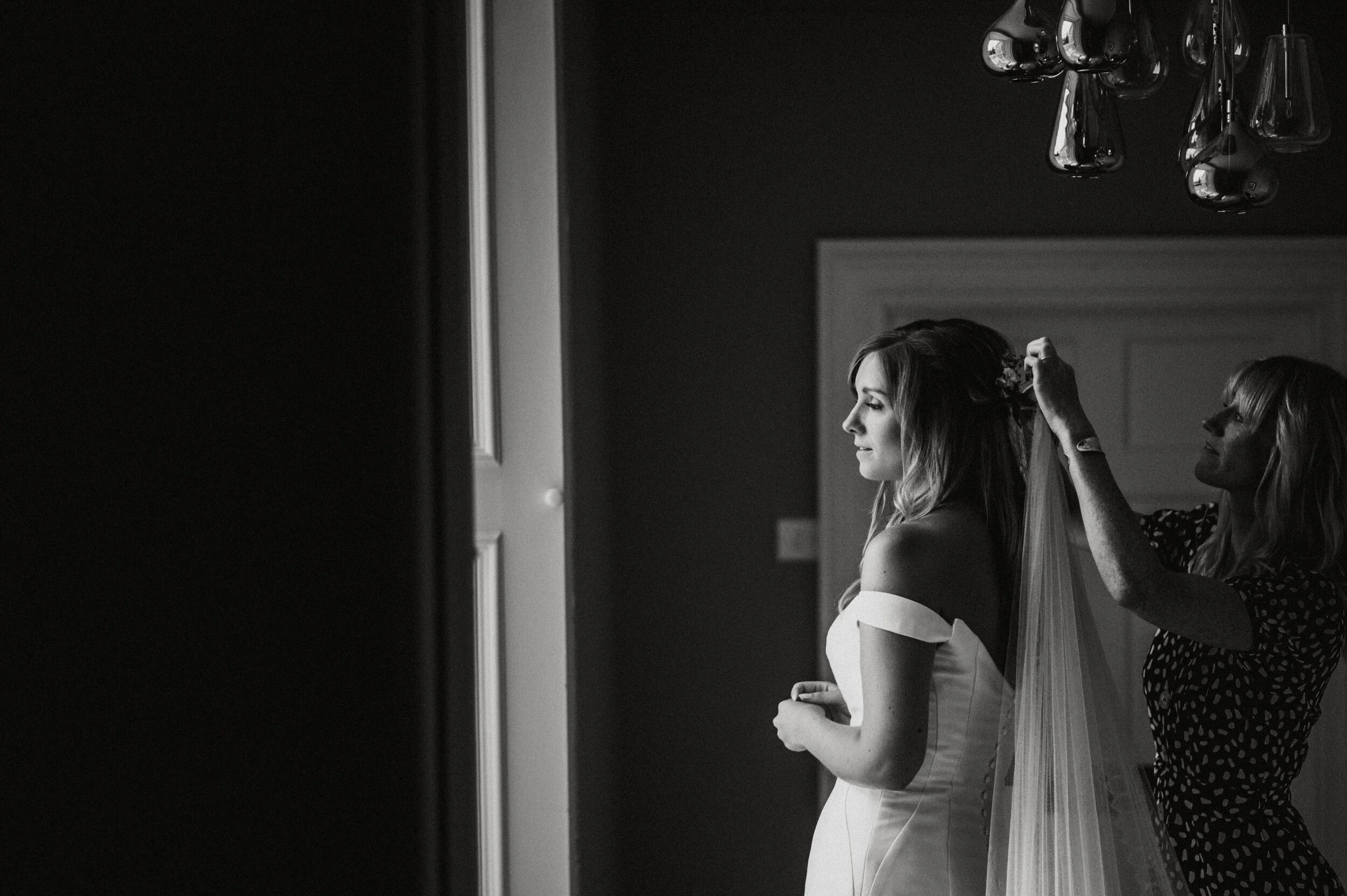 A bride getting ready, with a woman assisting her to put on a veil, as they stand near a window in a room.