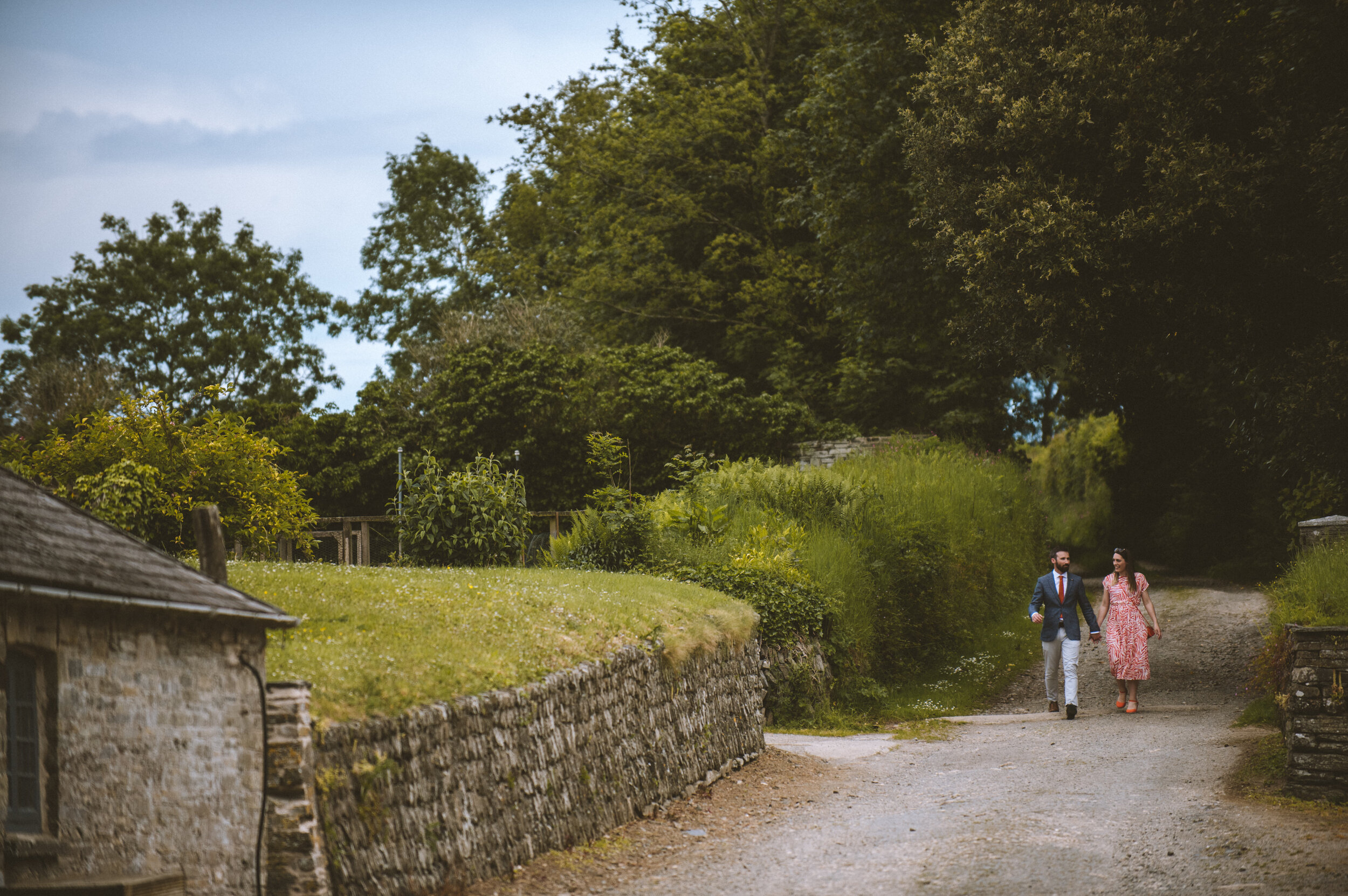 A man in a suit and a woman in a floral dress walking hand in hand along a gravel path in a rural area with greenery and trees.