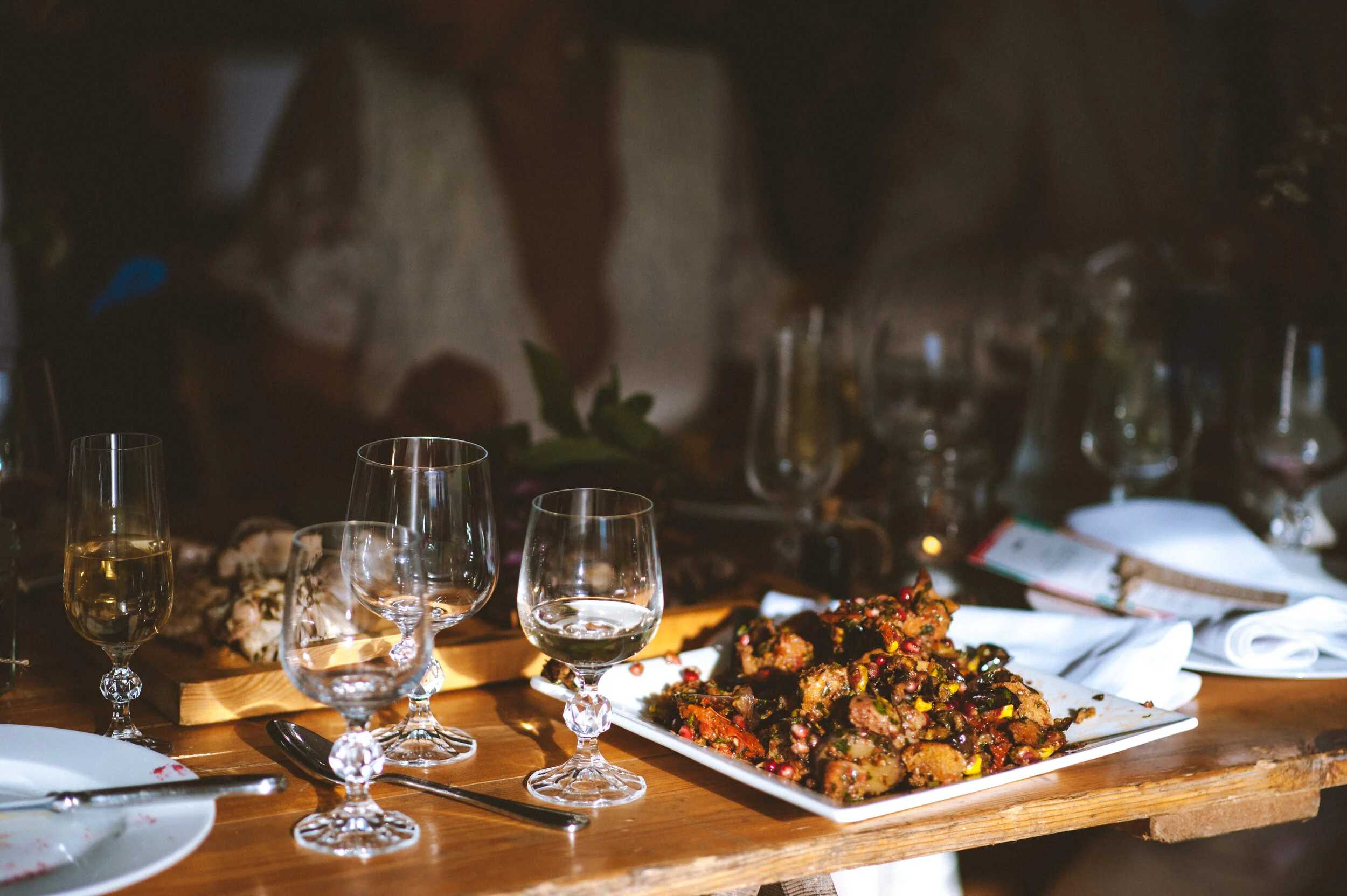 A rustic wooden table set with three empty whiskey glasses, a white square plate with a colorful meat and vegetable dish, empty white plates, silverware, and napkins. In the background, there are additional glasses, plates, and a cutting board.