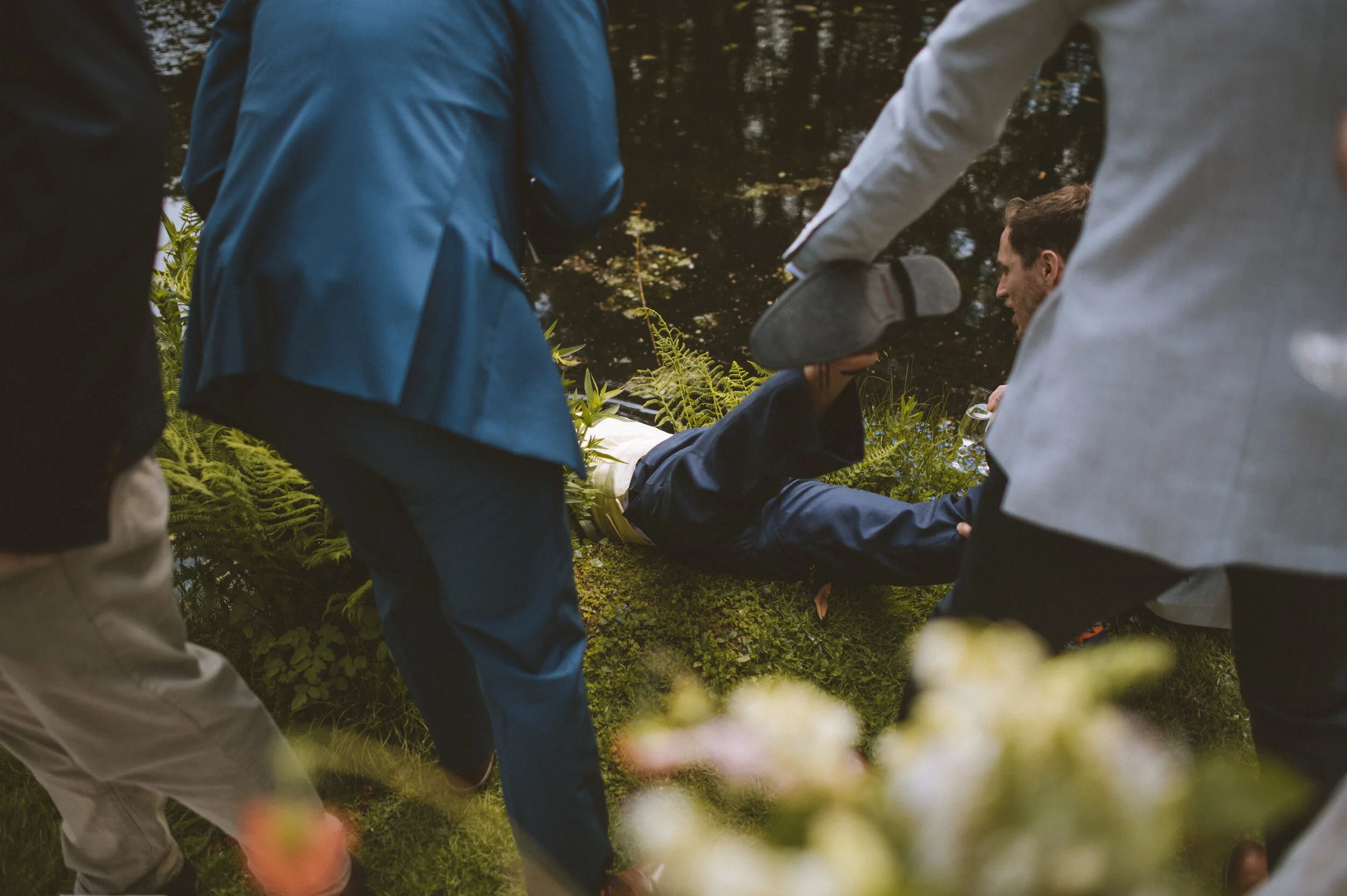 People surrounding a man lying on the grass near a pond, appearing to assist or attend to him during an outdoor event.