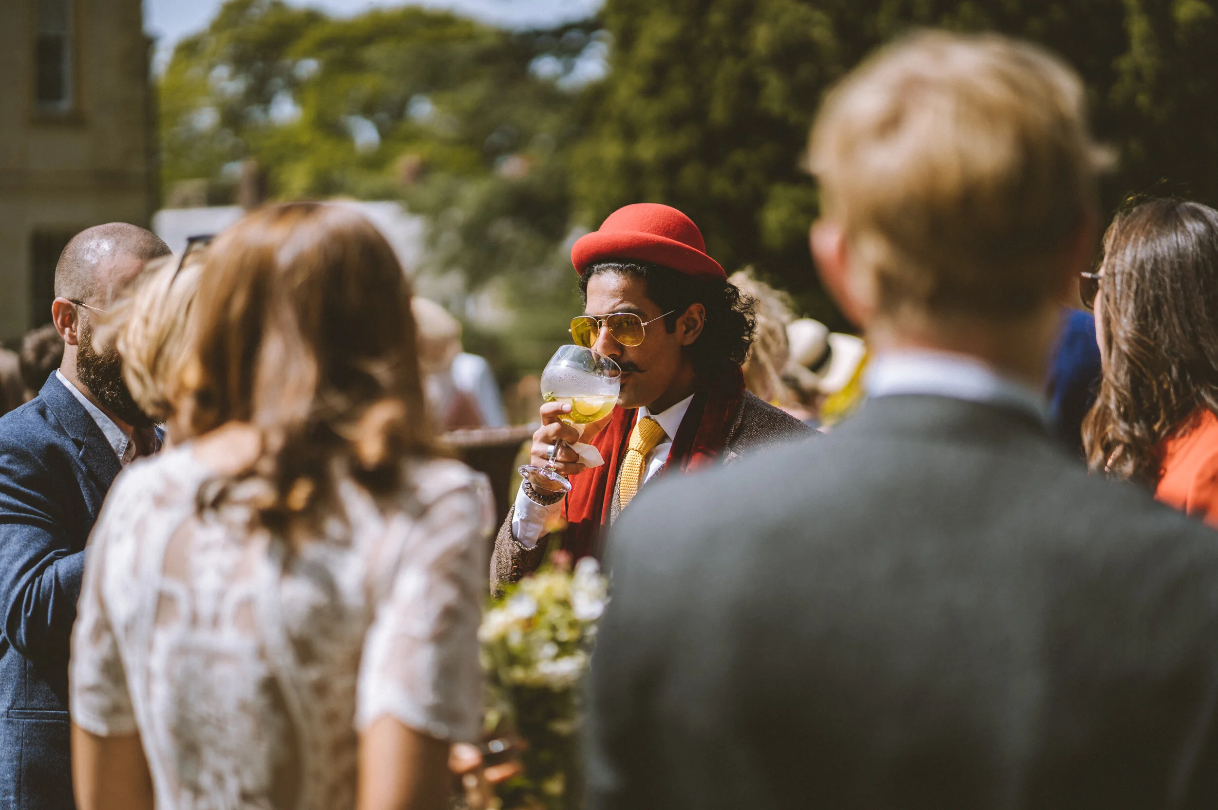 A man with curly hair, wearing sunglasses, a red hat, a yellow tie, and a brown jacket, is holding a cocktail glass and drinking outdoors at a social gathering.