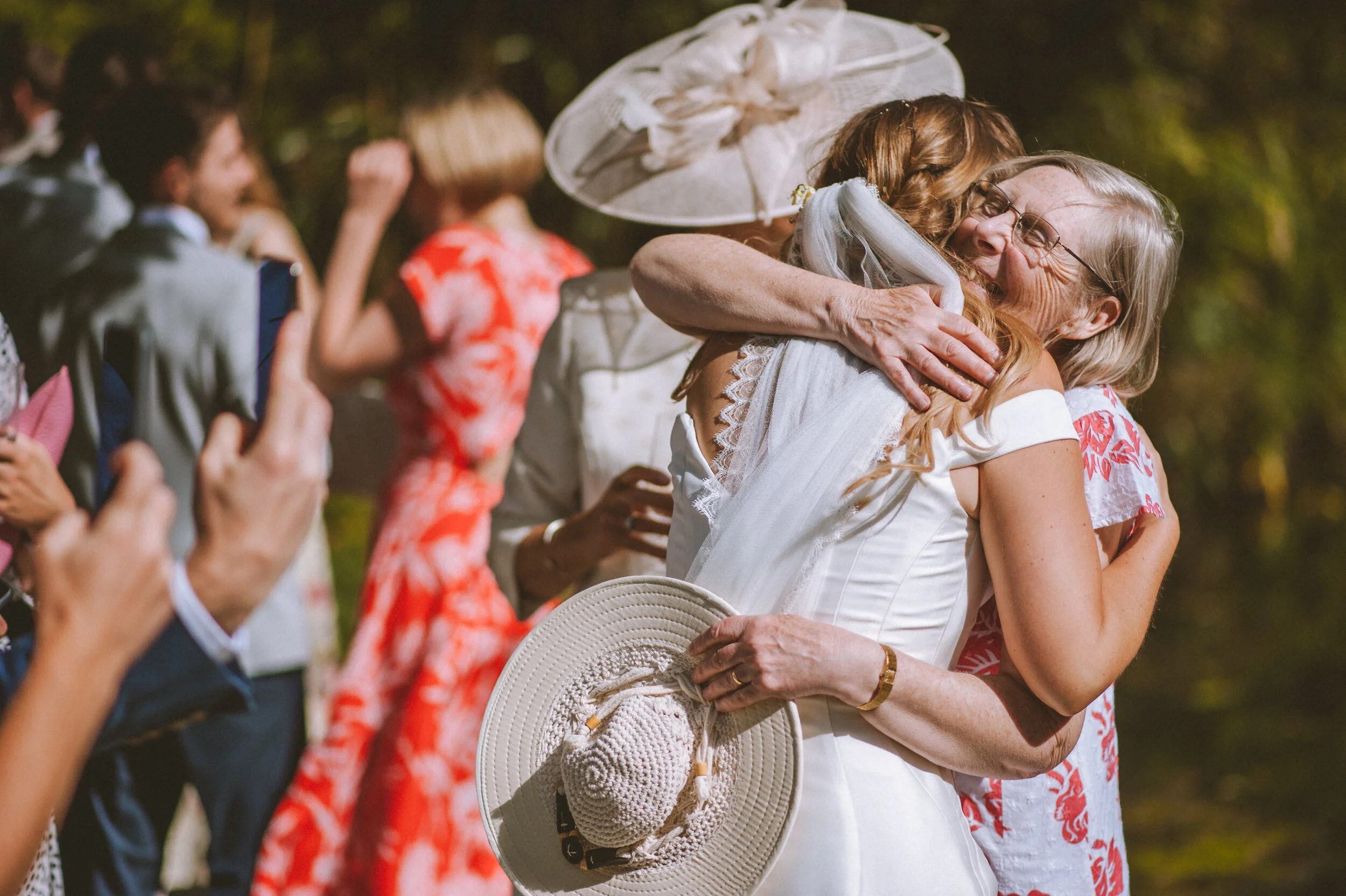 Two women hugging and smiling at a celebration outdoors, with people in the background taking photos and wearing colorful dresses.