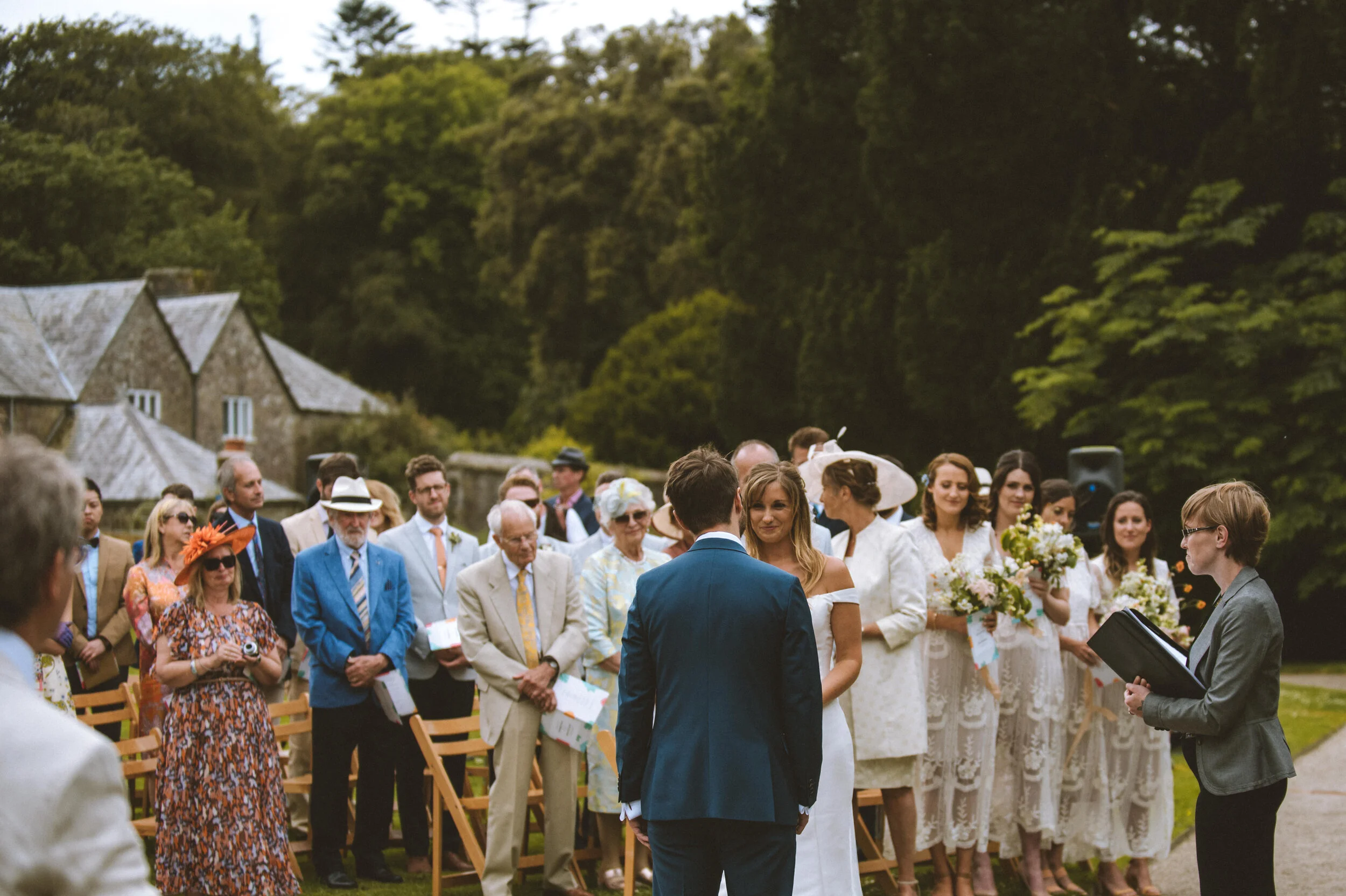 Wedding ceremony outdoors with the bride and groom facing each other, surrounded by guests, with lush green trees in the background.