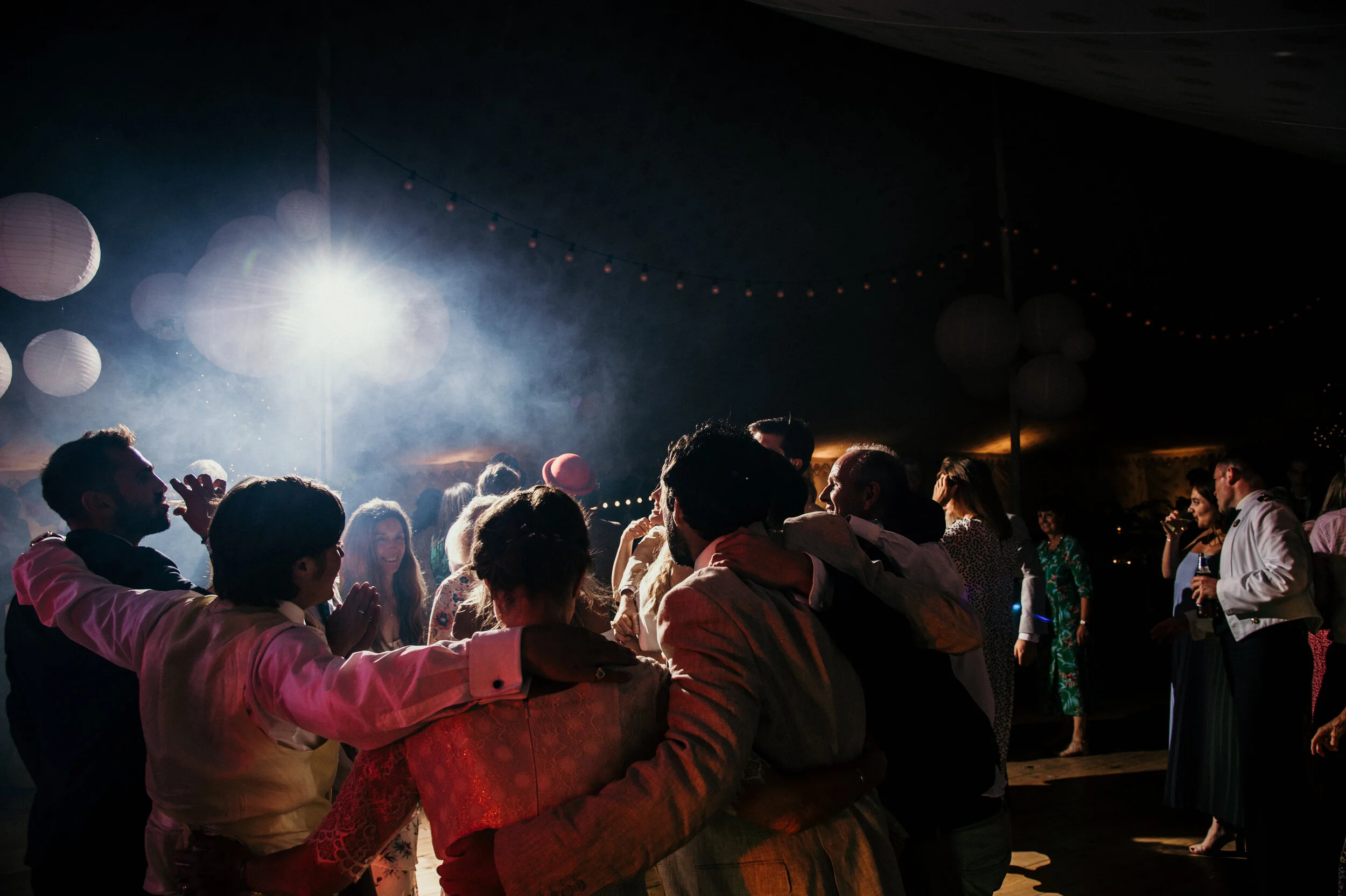 People dancing and socializing at a party in a dimly lit tent with paper lanterns and string lights, illuminated by a bright spotlight.