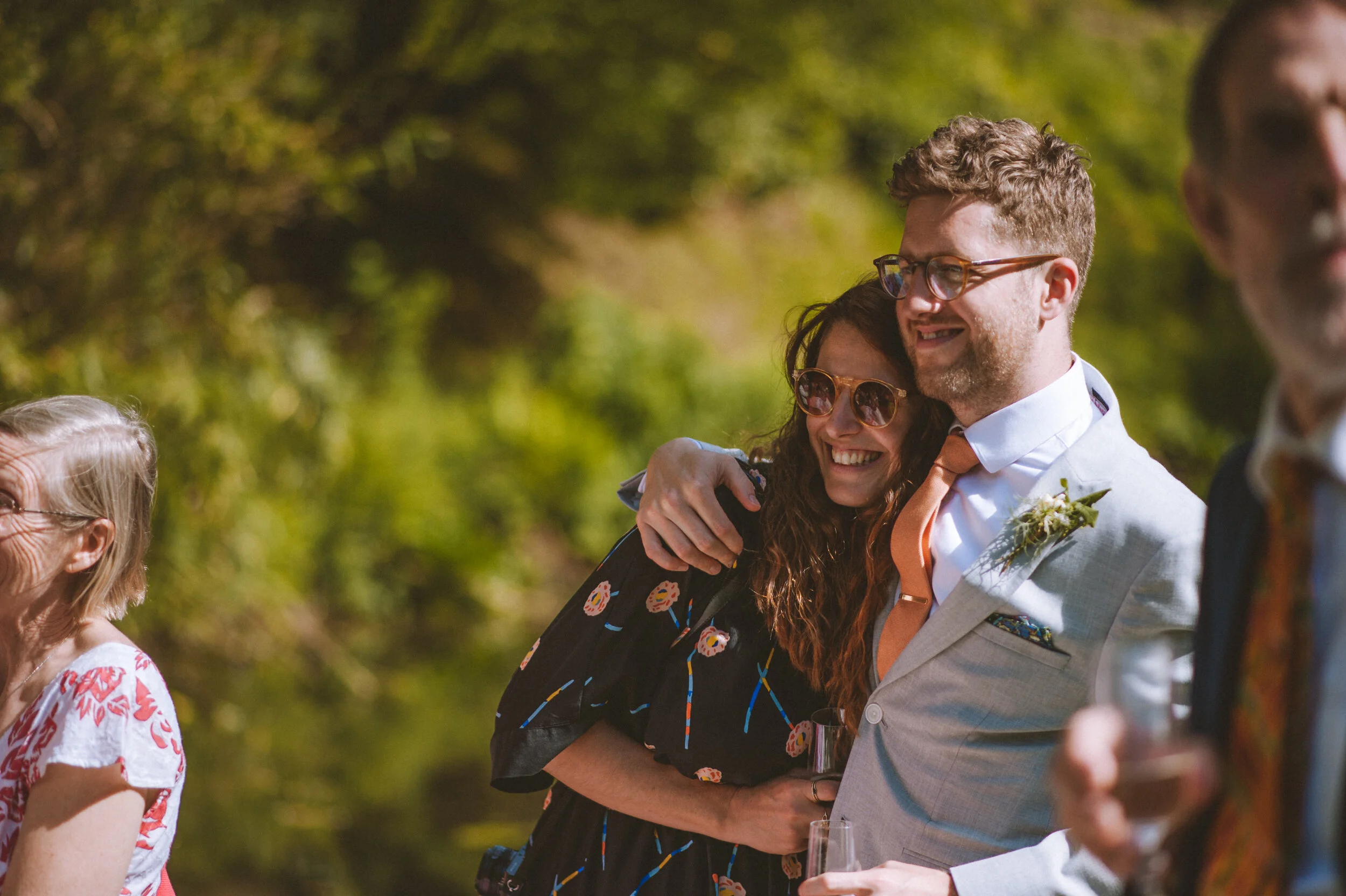 A group of people at an outdoor celebration, with a couple hugging in the center, smiling and wearing sunglasses, with trees in the background.