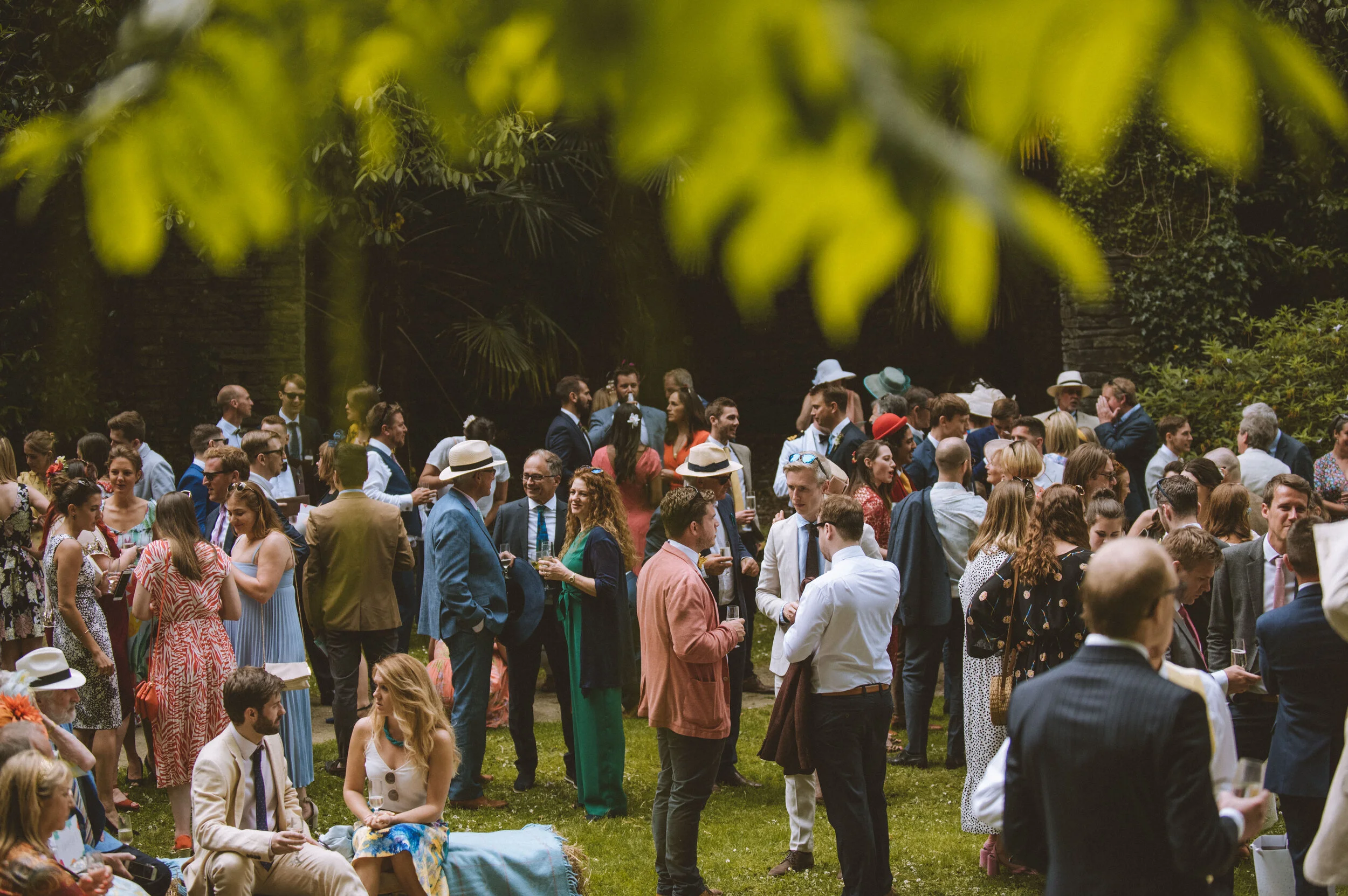 A large gathering of people at an outdoor event, dressed in semi-formal to formal attire, with some wearing hats, amidst lush greenery and trees.