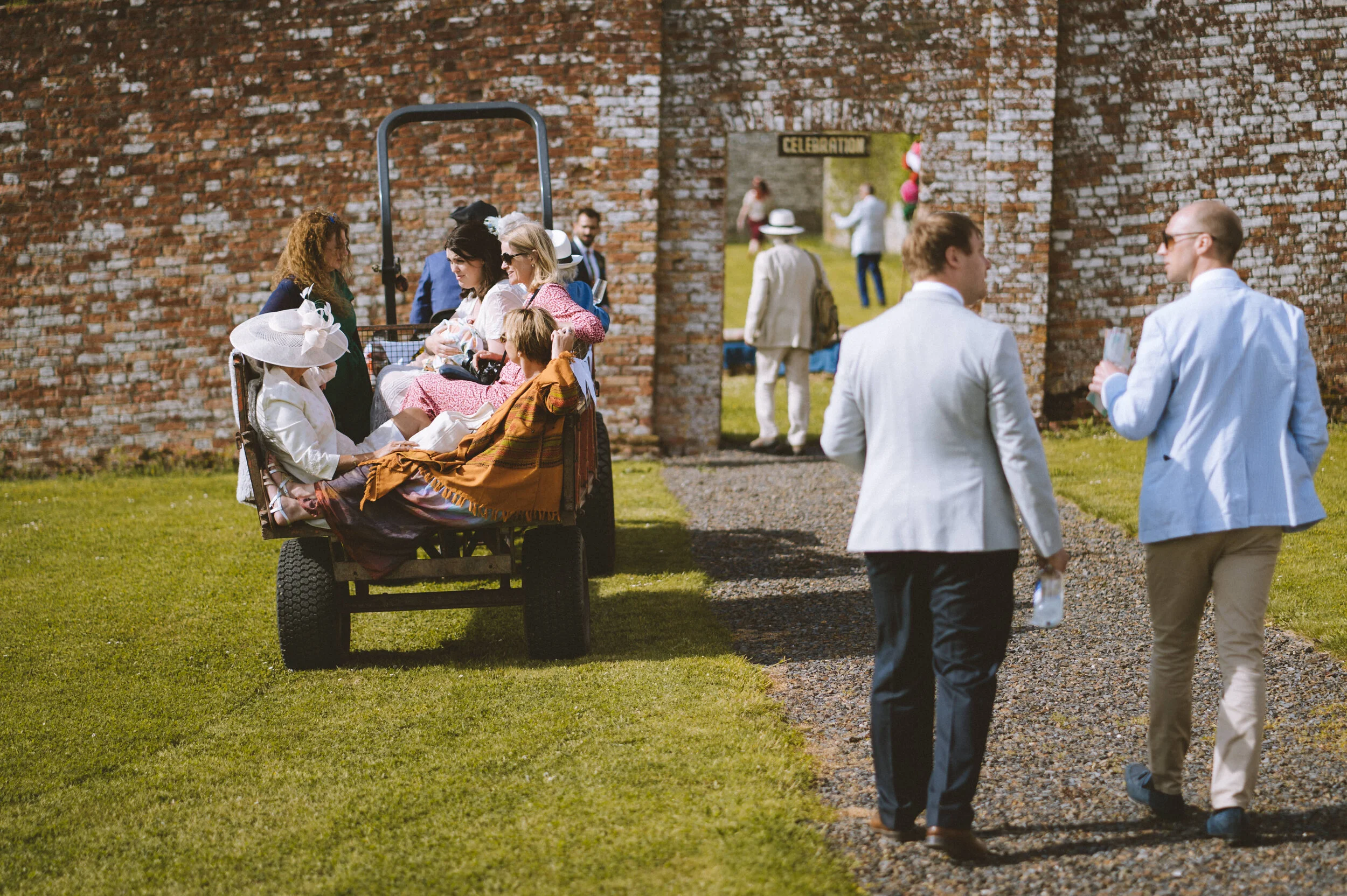 People gathering outdoors near a brick wall, with some sitting on a wagon and others standing or walking, at a daytime celebration event.