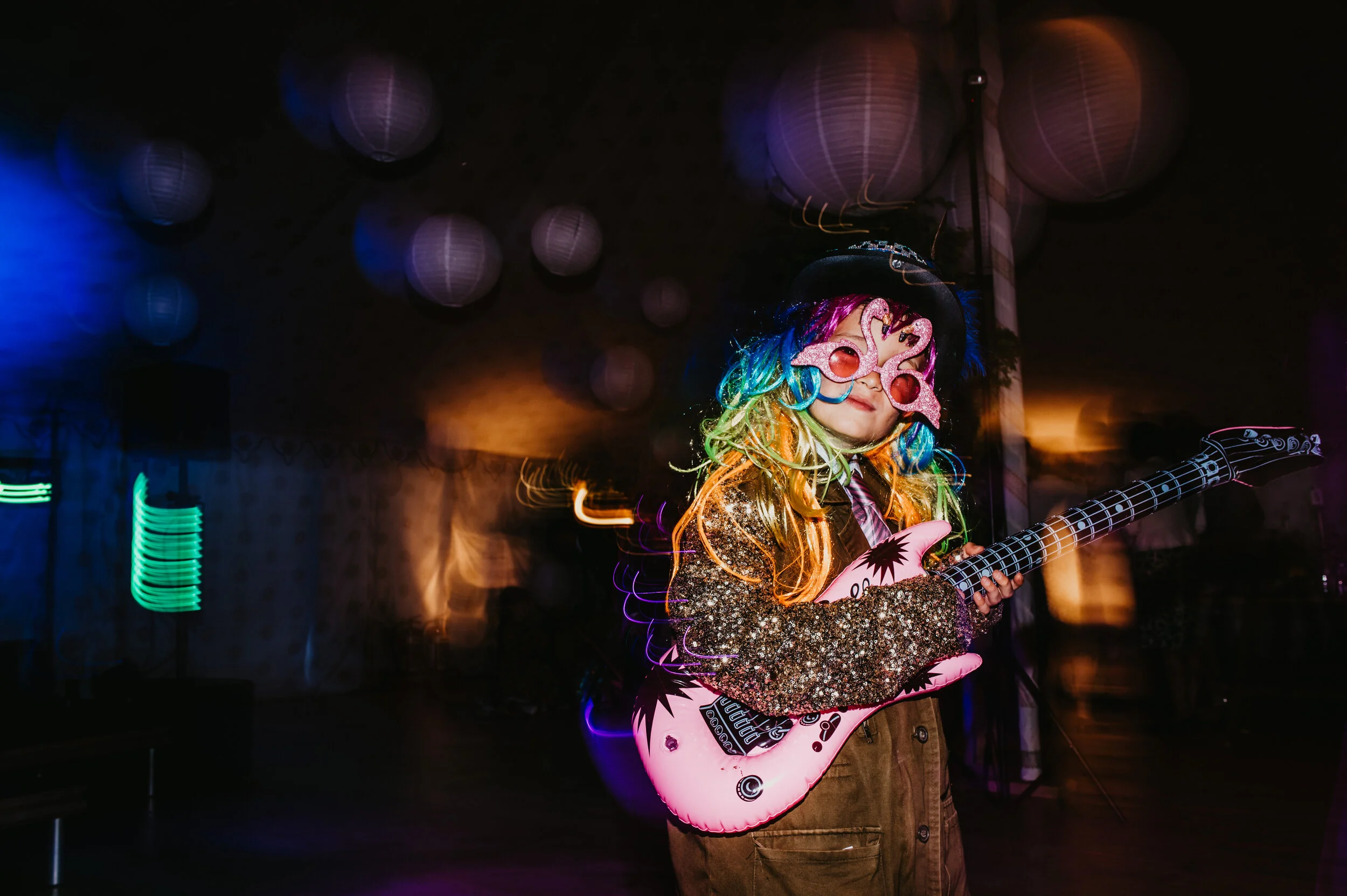 A young child dressed in a colorful, glittery costume with a rainbow wig and pink sunglasses, standing in a dimly lit room decorated with paper lanterns, holding an inflatable pink guitar.