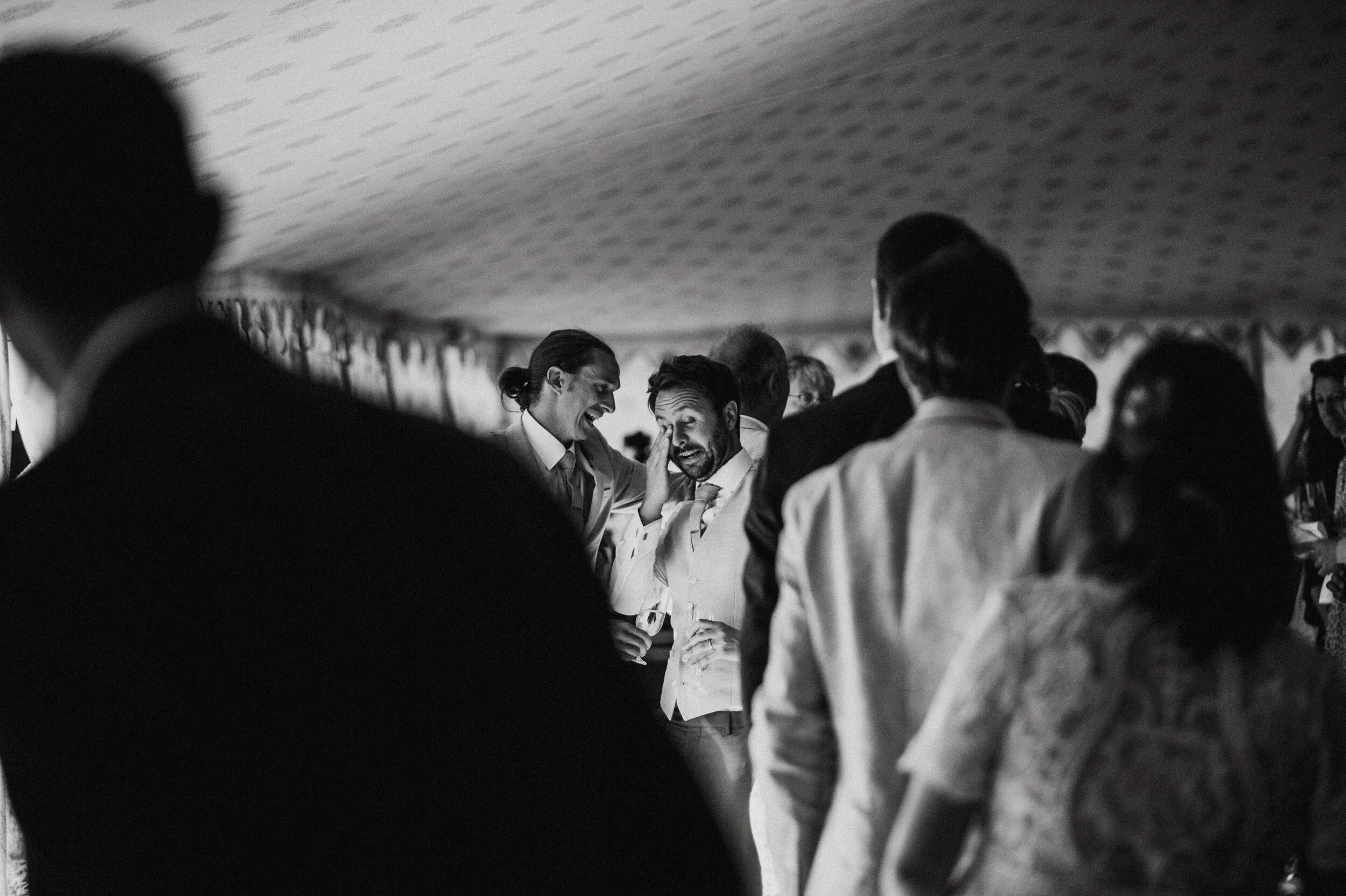 Black and white photo of people at a social gathering, with a man touching the face of another man who appears emotional. Several people are blurred in the foreground and background.