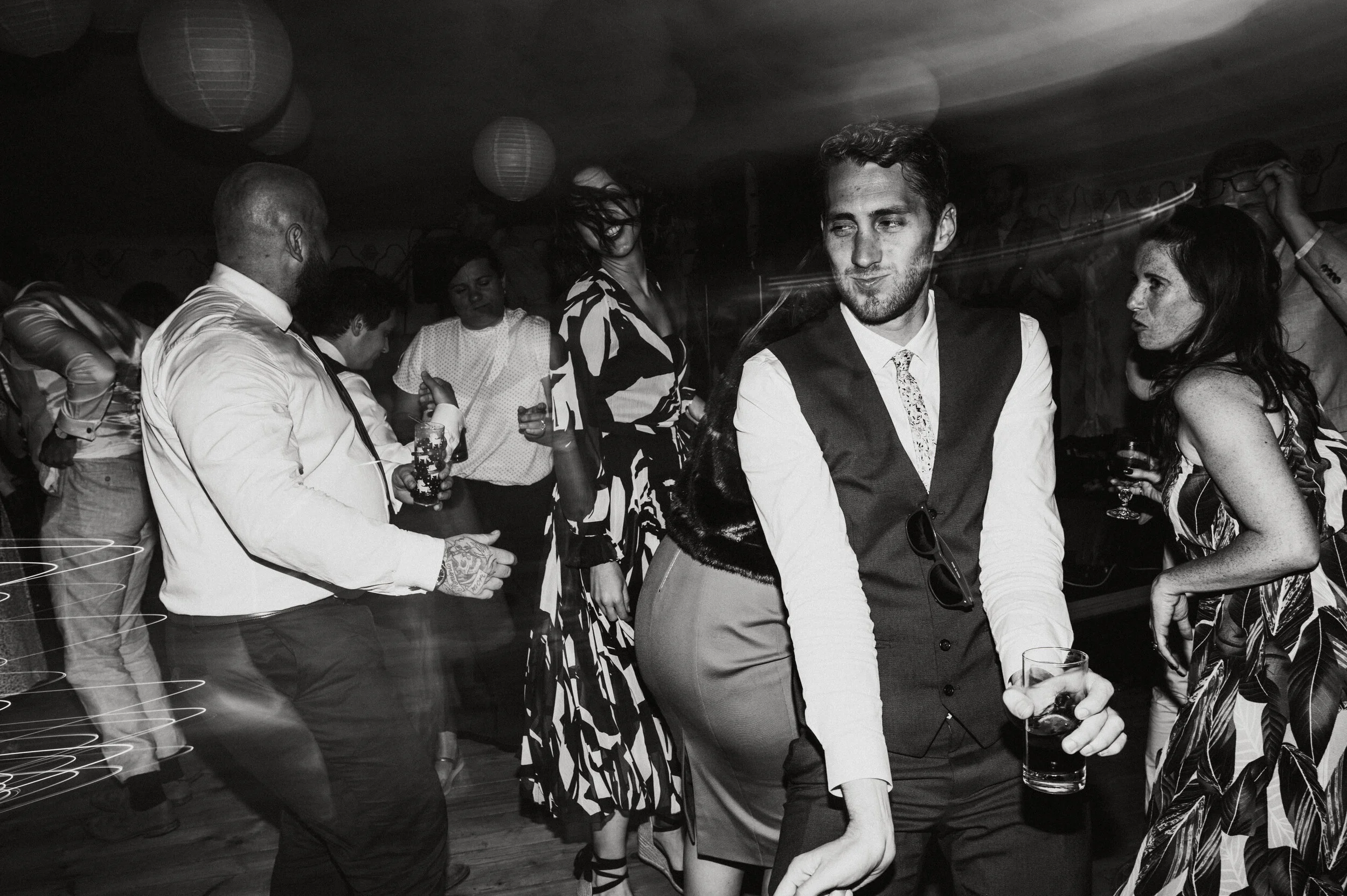 People dancing and socializing at a party or social gathering, black and white photo, some holding drinks, in an indoor setting with paper lanterns hanging from the ceiling.