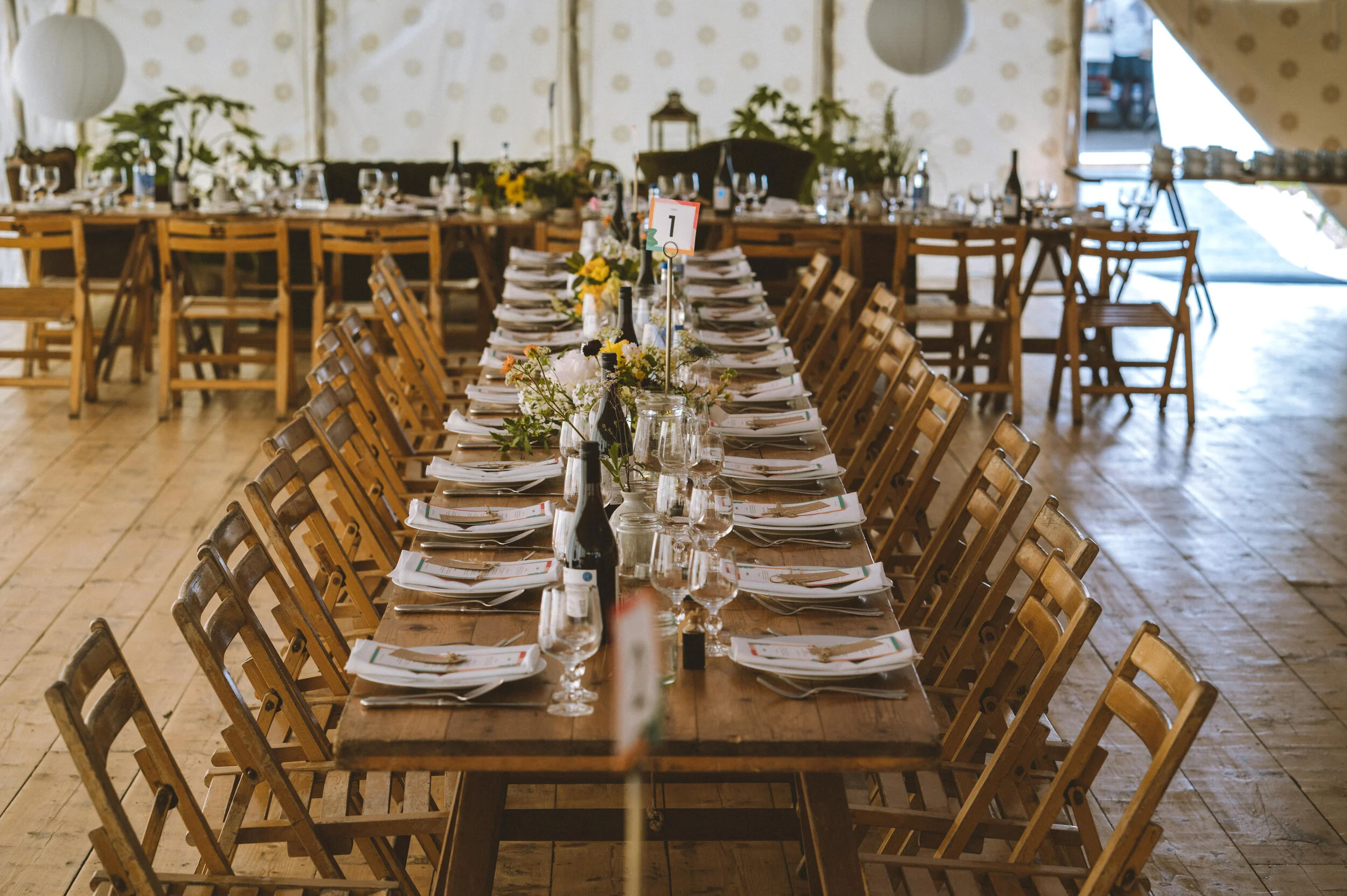 A long wooden banquet table set for a formal event, with chairs on both sides, adorned with flowers, wine bottles, glasses, and place settings, in a decorated indoor space.