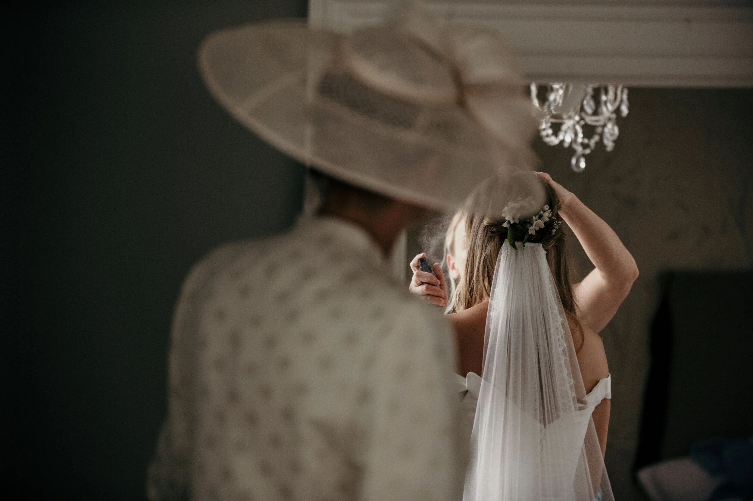 A woman with a veil and floral headpiece adjusts her hair while another woman in a wide-brimmed hat prepares herself for a wedding or special occasion.