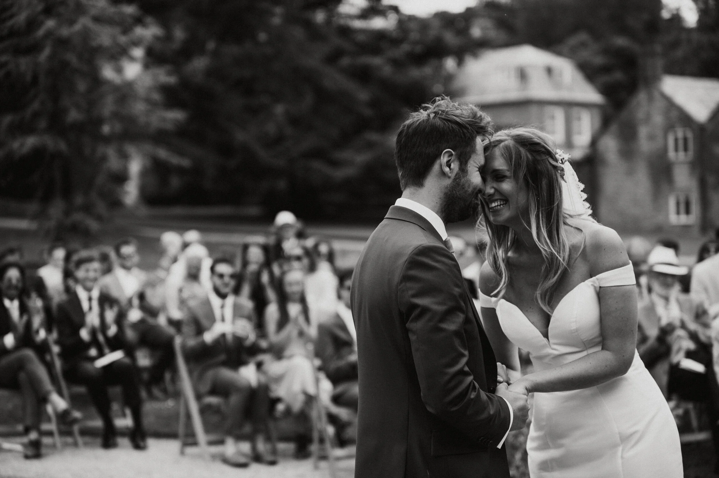 A black and white photograph of a bride and groom smiling and touching foreheads during their outdoor wedding ceremony, with guests seated in the background.