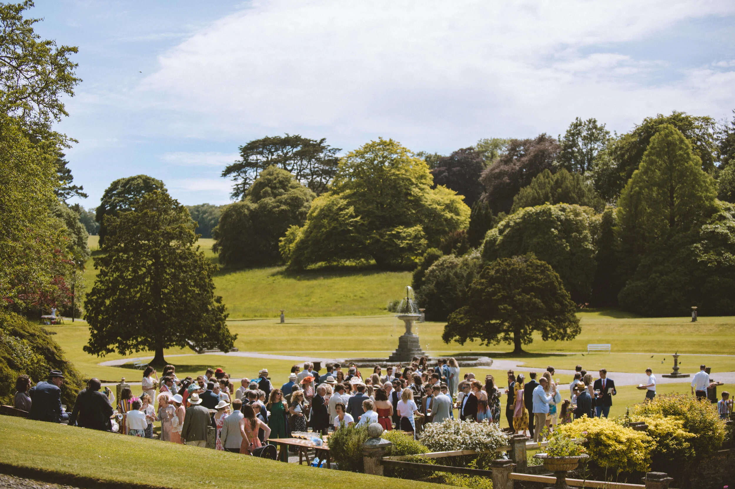 Pencarrow House wedding photography in Cornwall - A large outdoor gathering of people in formal attire in a park with a fountain, surrounded by lush trees and green grass under a partly cloudy sky.