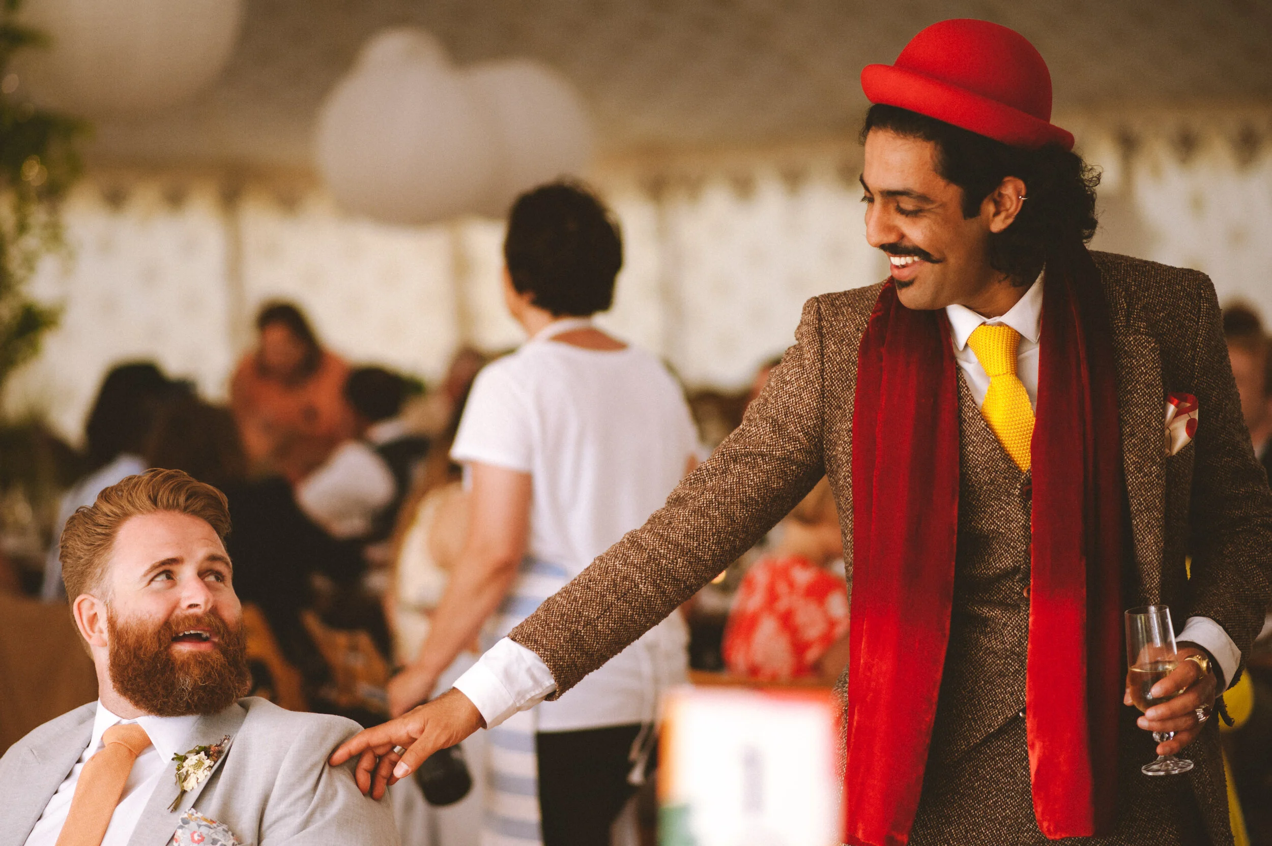 A man with a red hat, brown suit, yellow tie, and red scarf is smiling and touching a man with a beard, gray suit, and orange tie at a gathering or party.