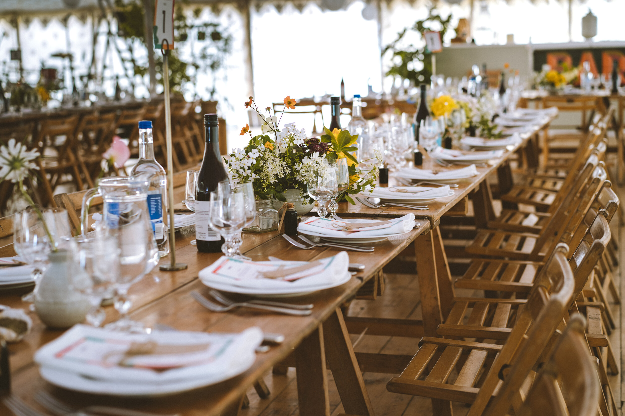 Long wooden banquet table set with plates, silverware, wine glasses, bottles of wine and water, and floral centerpieces, in a well-lit outdoor venue with wooden chairs.