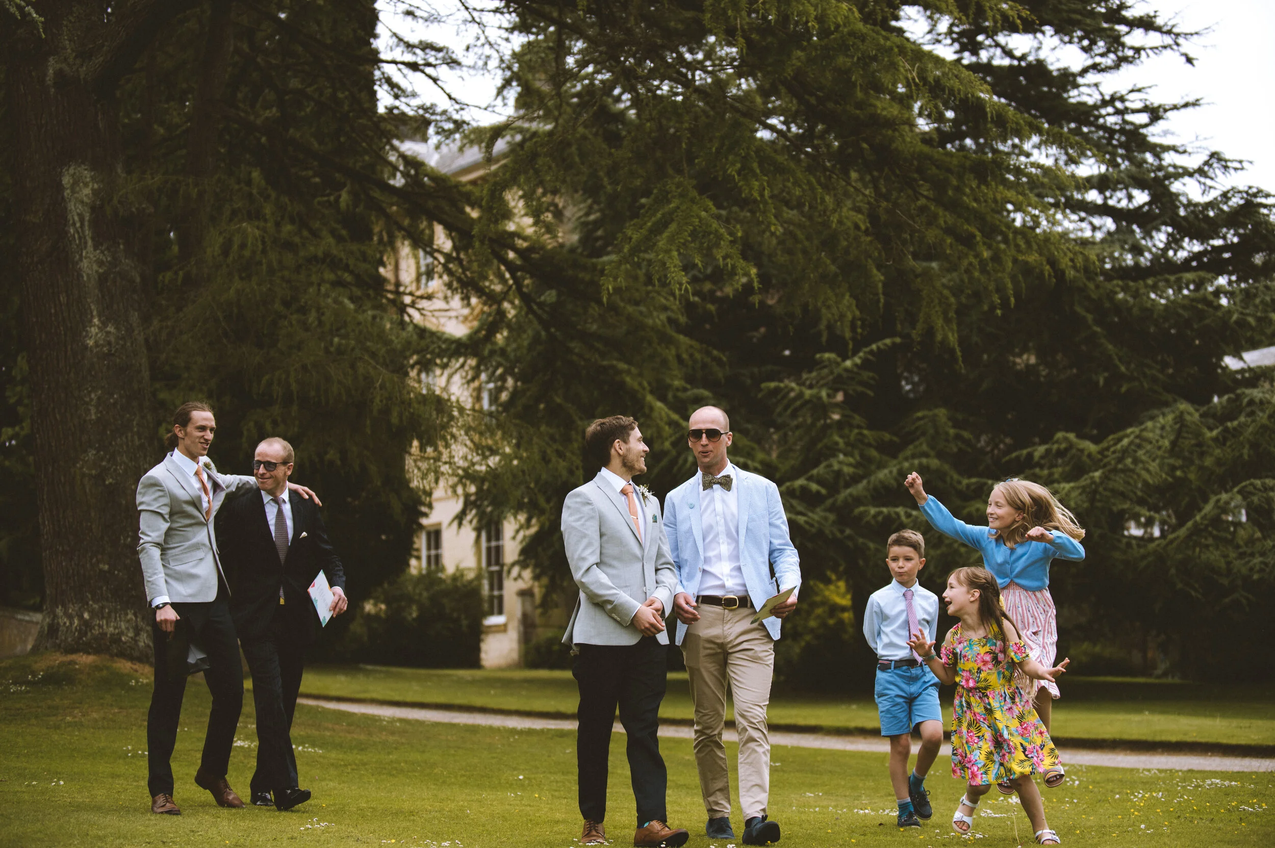 Group of people walking and playing on a grassy lawn with large trees and a building in the background.
