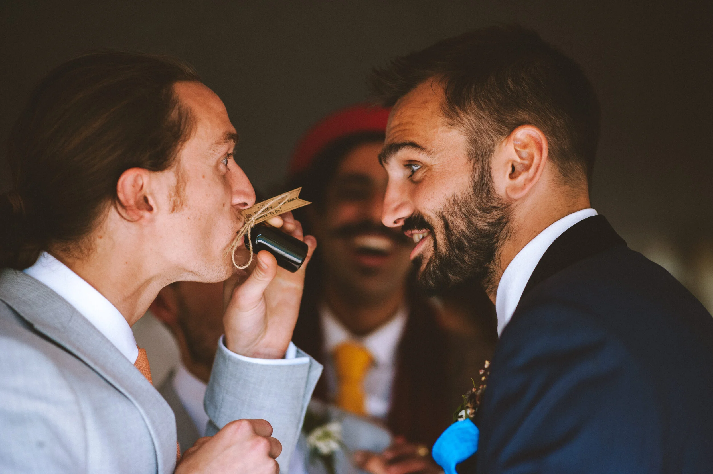 Two men in formal suits face to face, about to kiss while one holds a small black object with a string tied to it near the other's lips, with a third person smiling in the background.