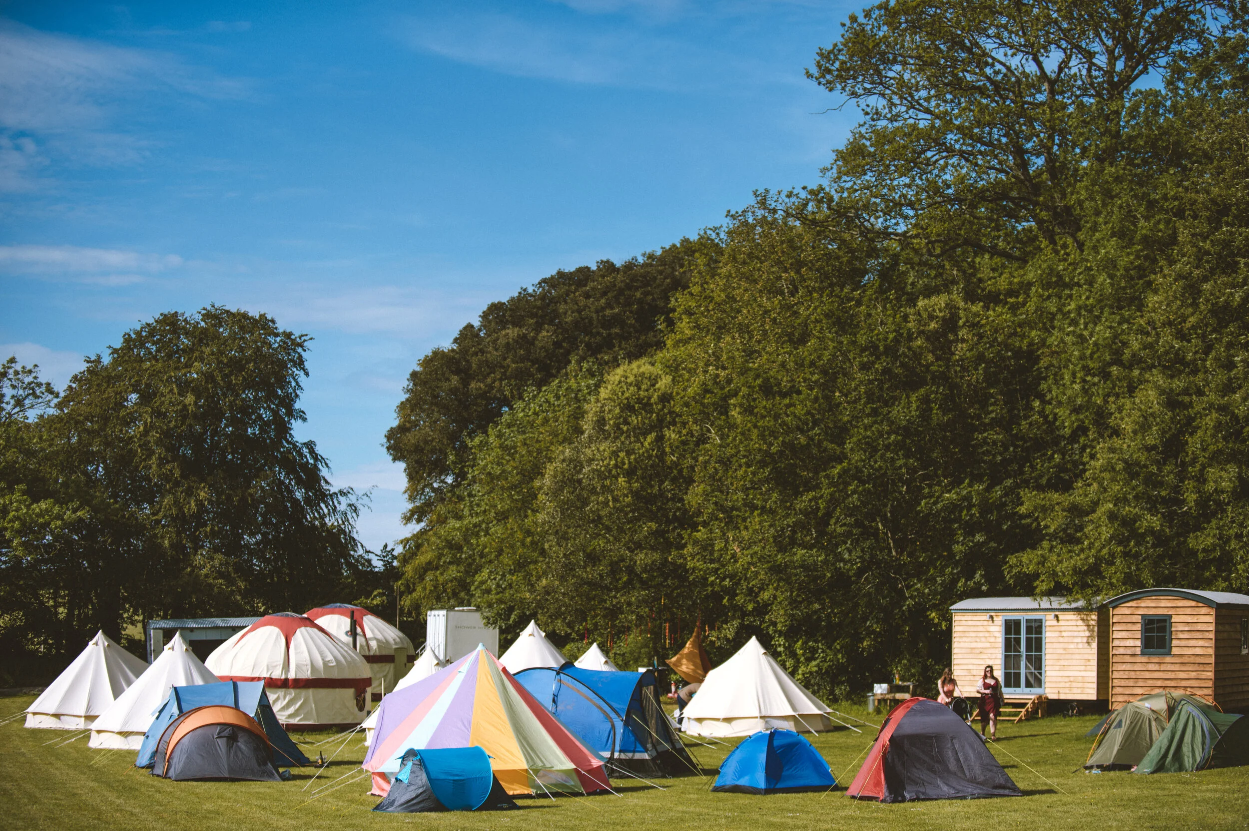 Multiple tents of various colors set up on a grassy field, with large green trees and a blue sky in the background. Two people are walking near a small wooden building.