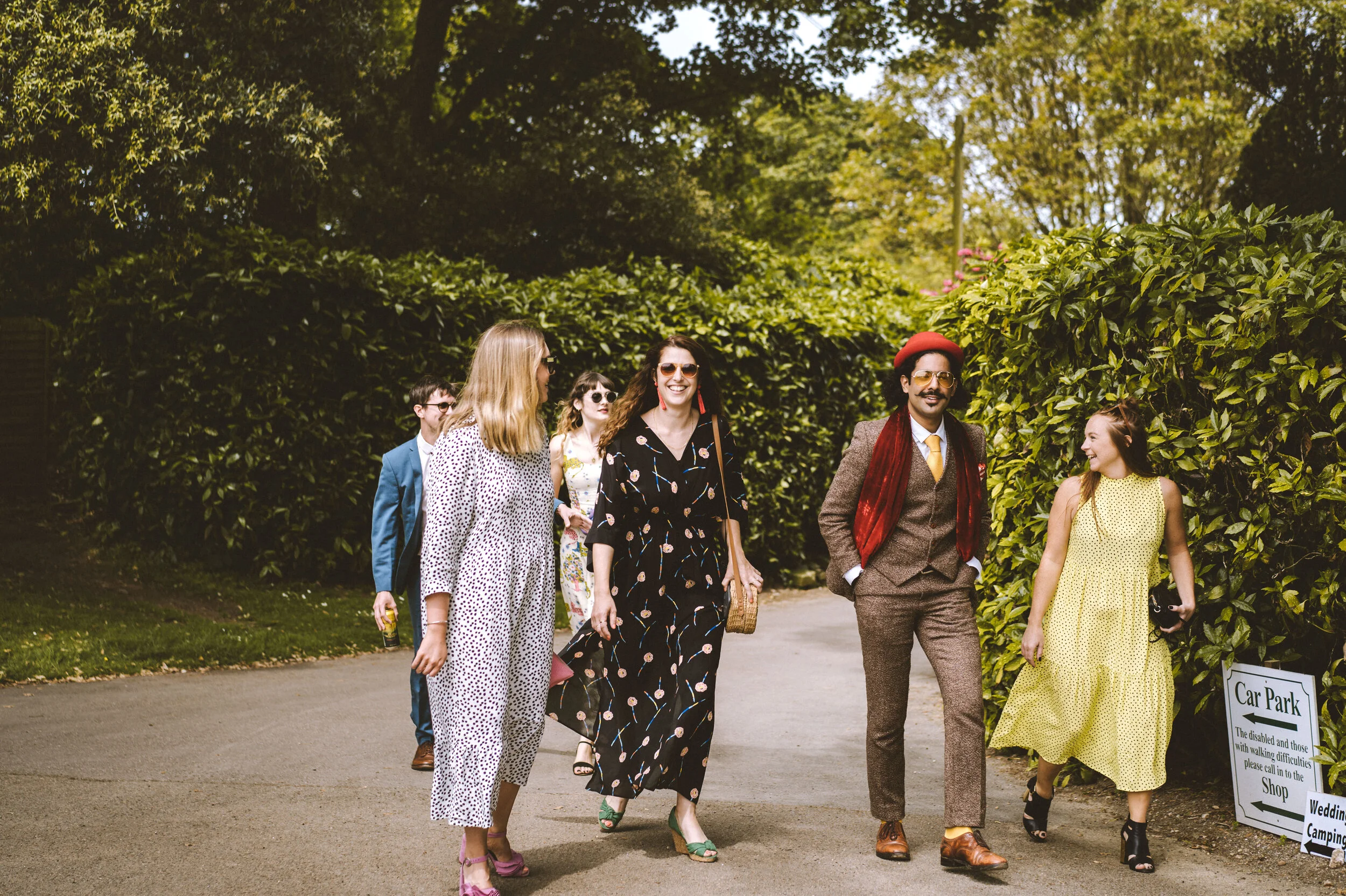 A group of six people walking along a park pathway, surrounded by green trees and bushes, with a sign indicating a car park and directions for wedding and camping.