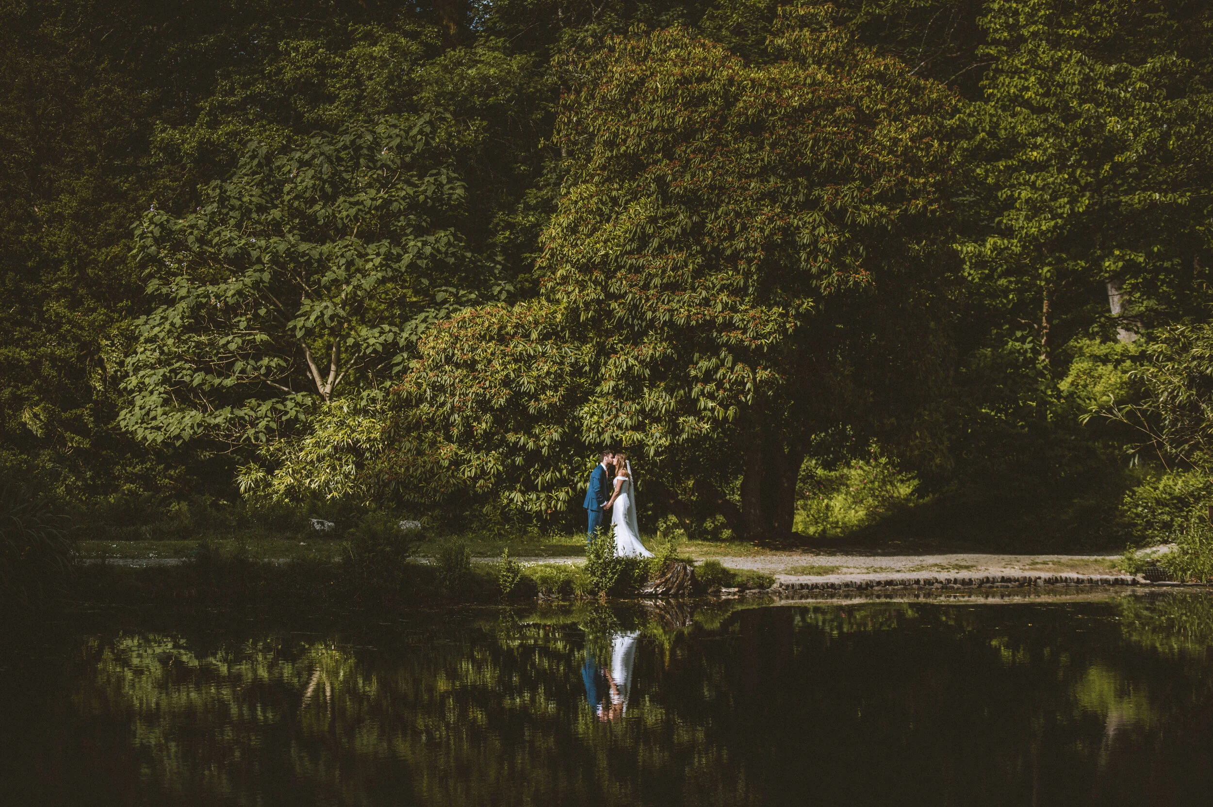 A bride and groom holding hands by a pond in a wooded area during daytime.