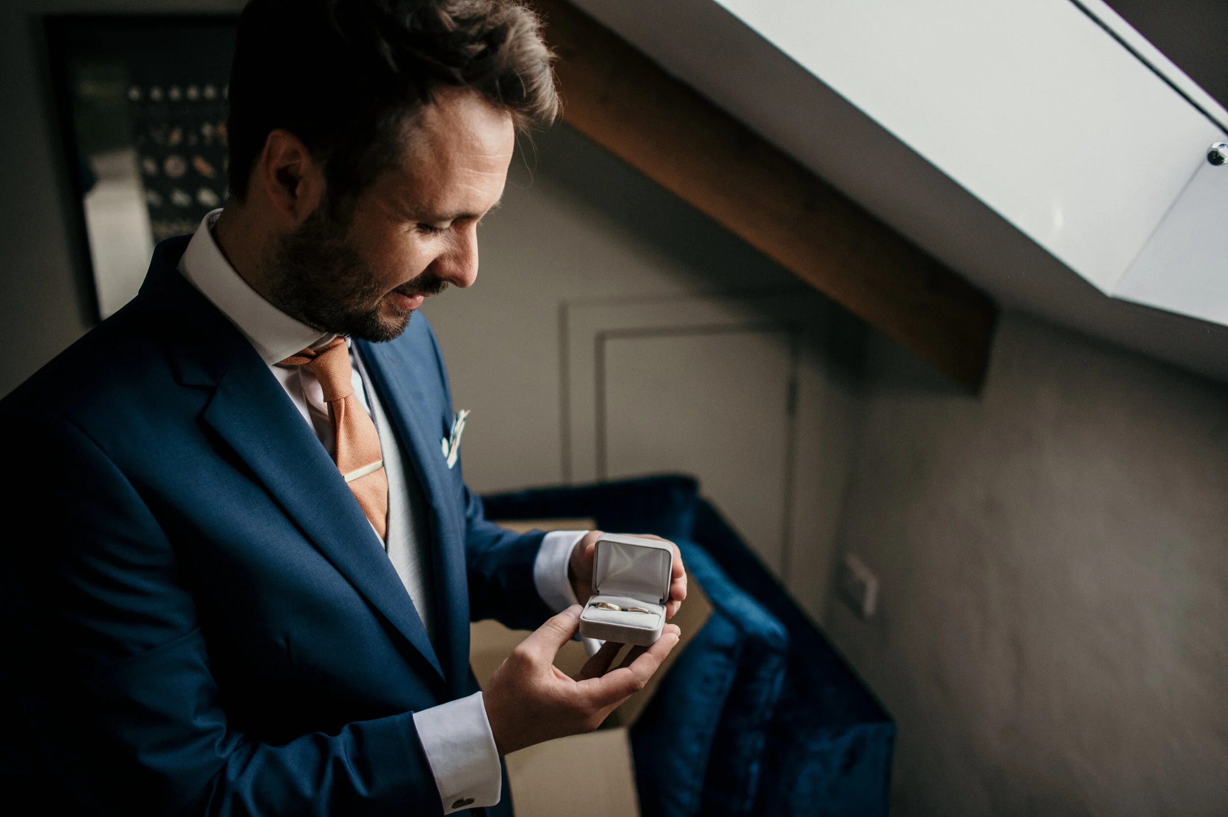 A man in a blue suit and white shirt, holding an open ring box with a wedding ring, looking at it with a smile.