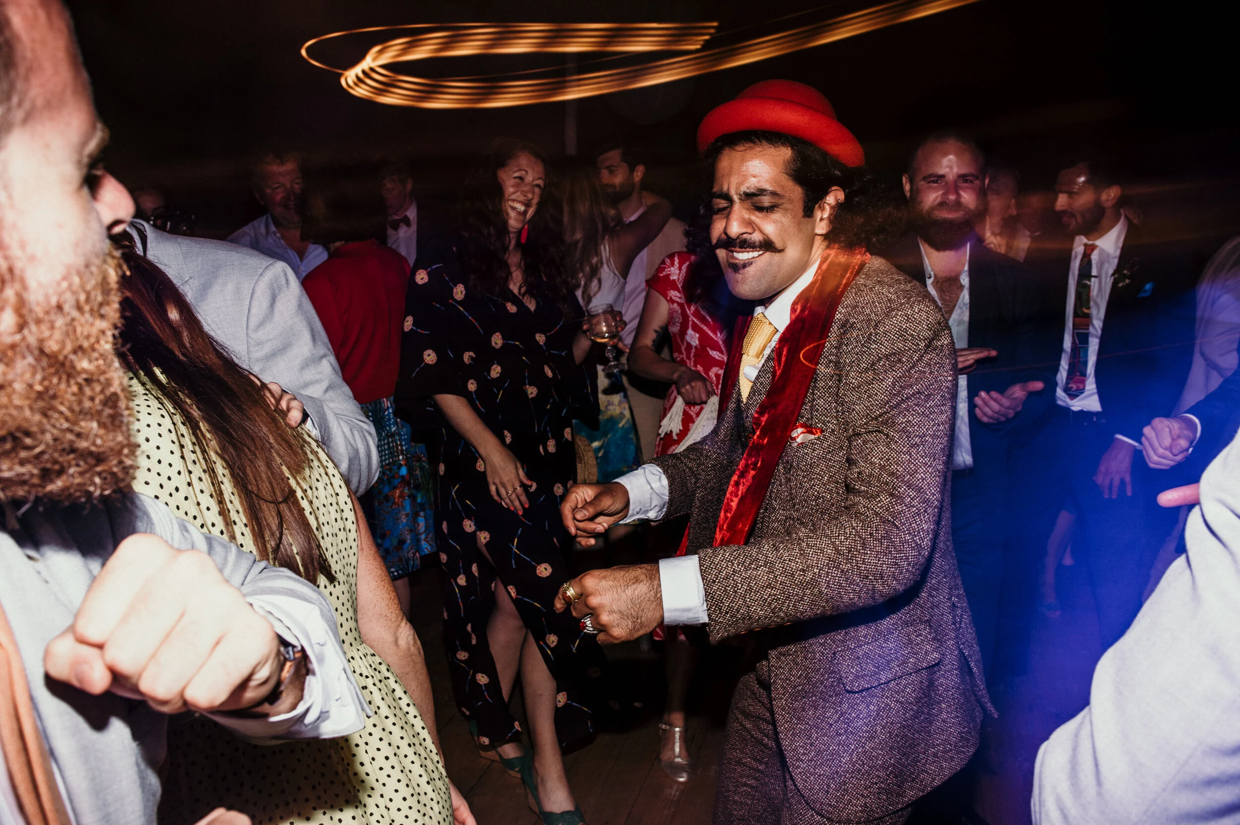 People dancing and socializing at a lively party or celebration, with a man in a brown suit, red hat, and a shiny red sash dancing in the center.