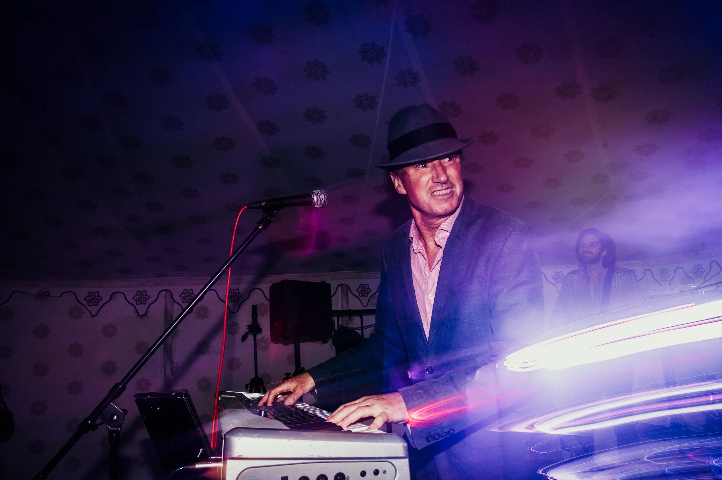 A man wearing a fedora hat and suit jacket playing a keyboard on stage with colorful light streaks and a smiling man in the background.