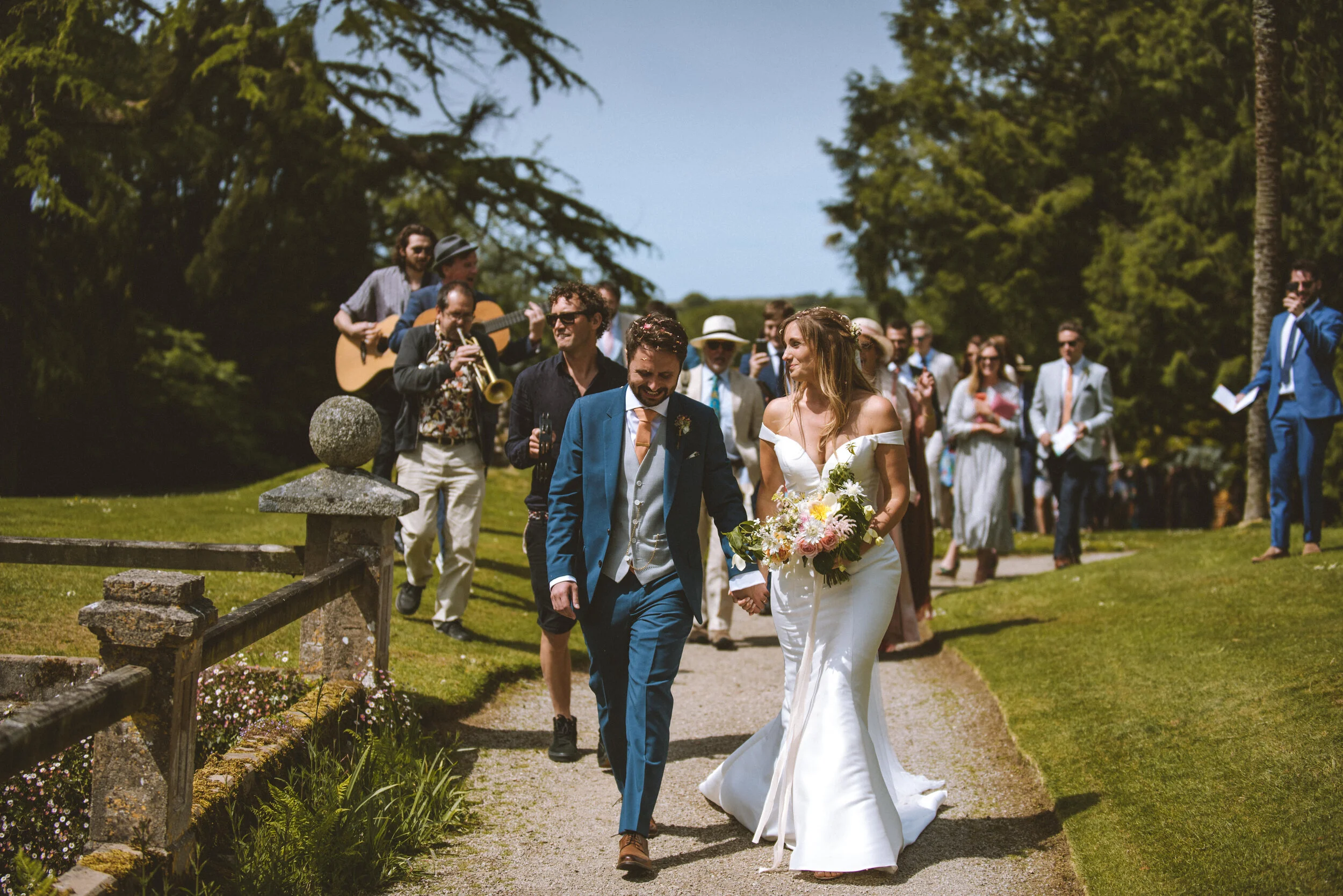 A bride and groom walking hand in hand outdoors during a wedding ceremony, surrounded by guests and musicians, with trees in the background.