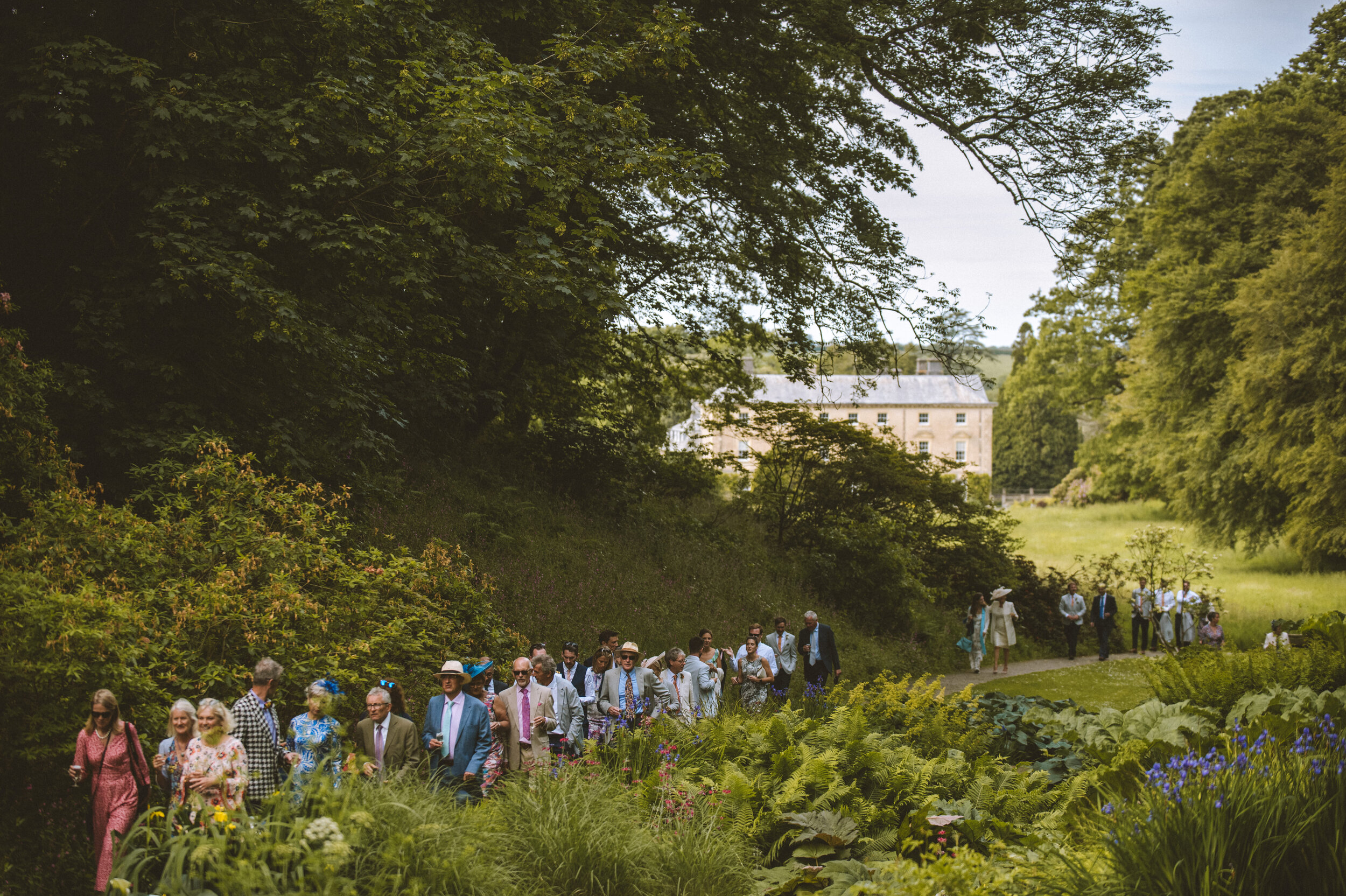 Group of well-dressed people walking through a lush green garden with trees and a large historic building in the background.