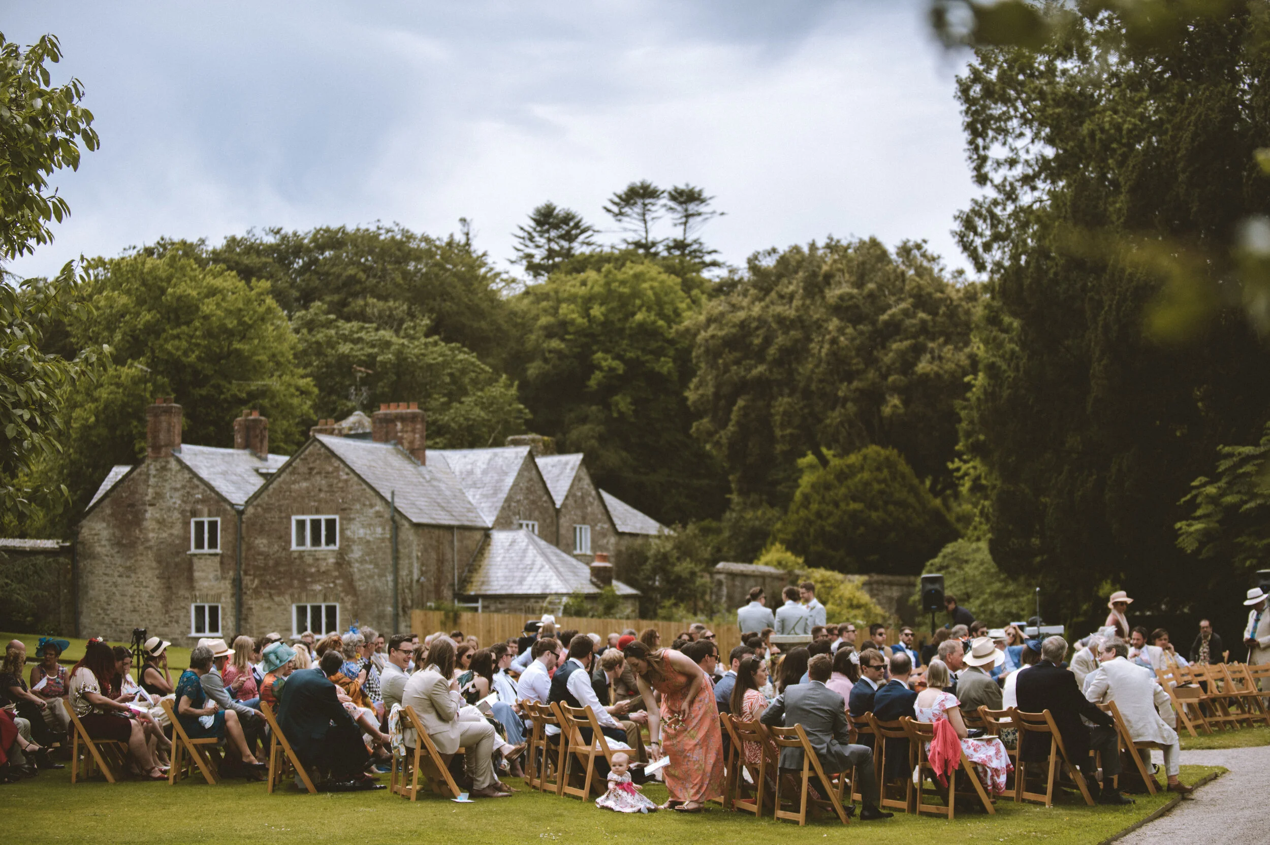 An outdoor wedding ceremony with guests seated on wooden chairs on a grassy area, surrounded by trees and brick buildings in the background, under a partly cloudy sky.