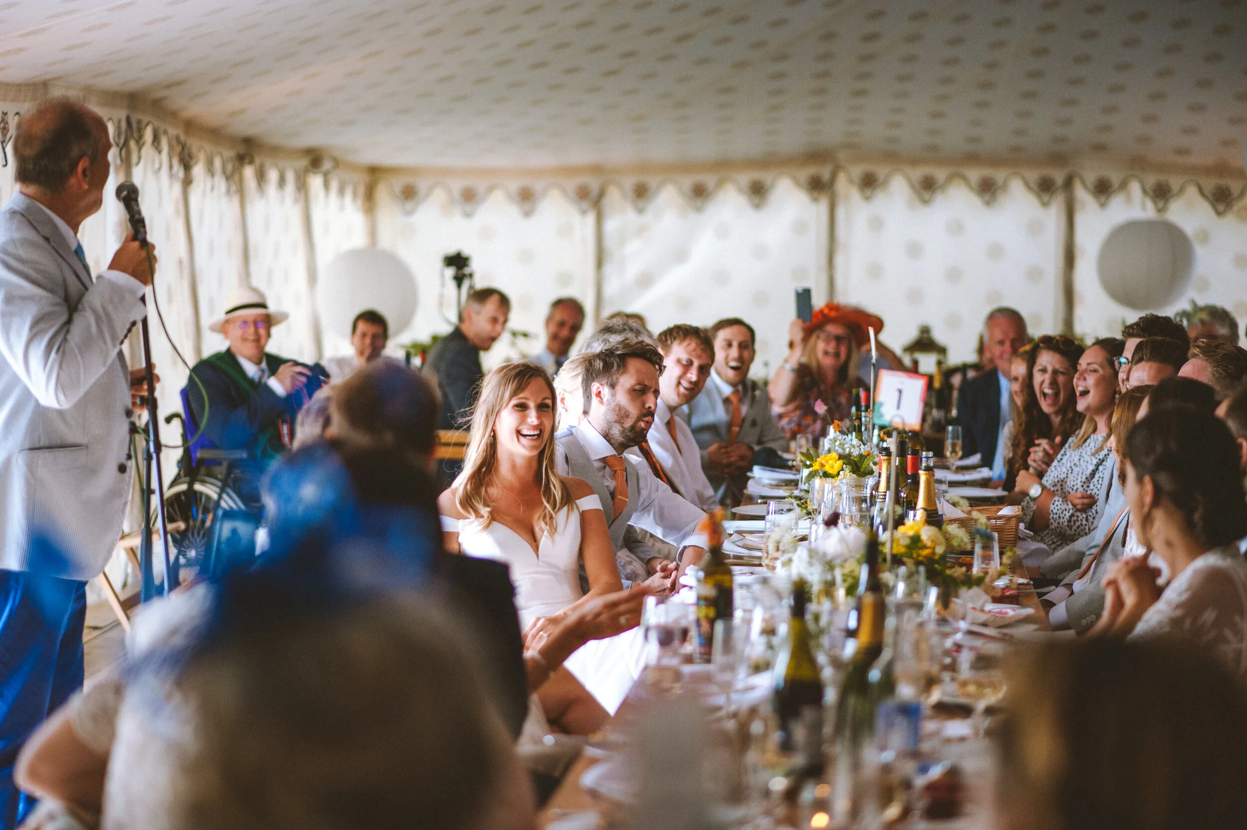 People seated at a long banquet table inside a decorated tent, listening to a man in a light-colored suit giving a speech, with guests dressed in formal attire, laughing and smiling.