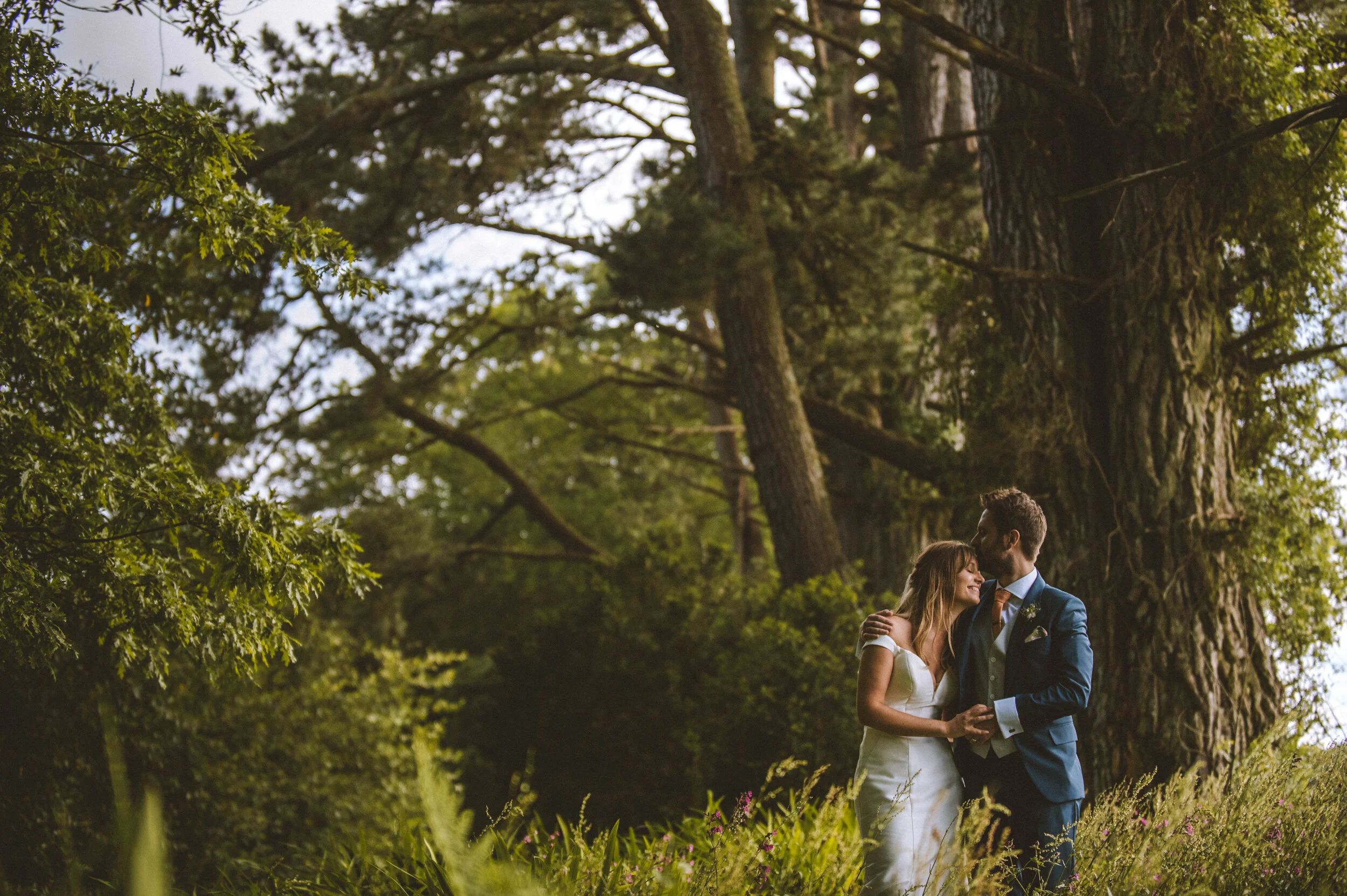 A bride and groom standing close together outdoors next to a large tree, holding hands and smiling at each other during their wedding.