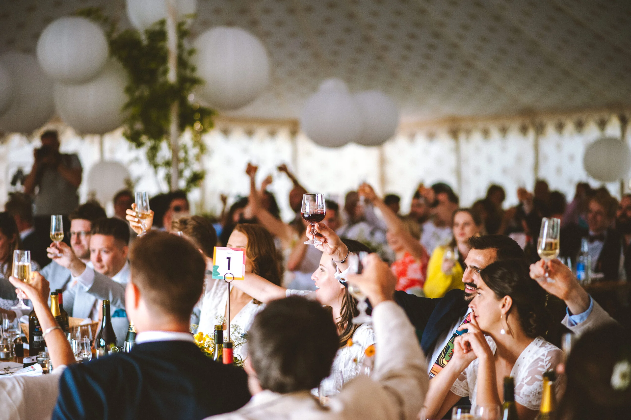 A large group of people at a celebration in a decorated tent, raising glasses of champagne and wine for a toast.