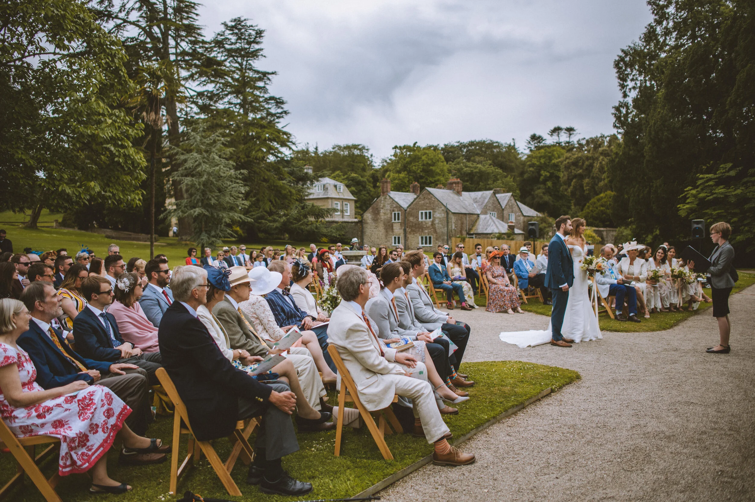 Outdoor wedding ceremony with bride and groom standing before officiant, seated guests on both sides, lush green trees, and historic stone buildings in the background.