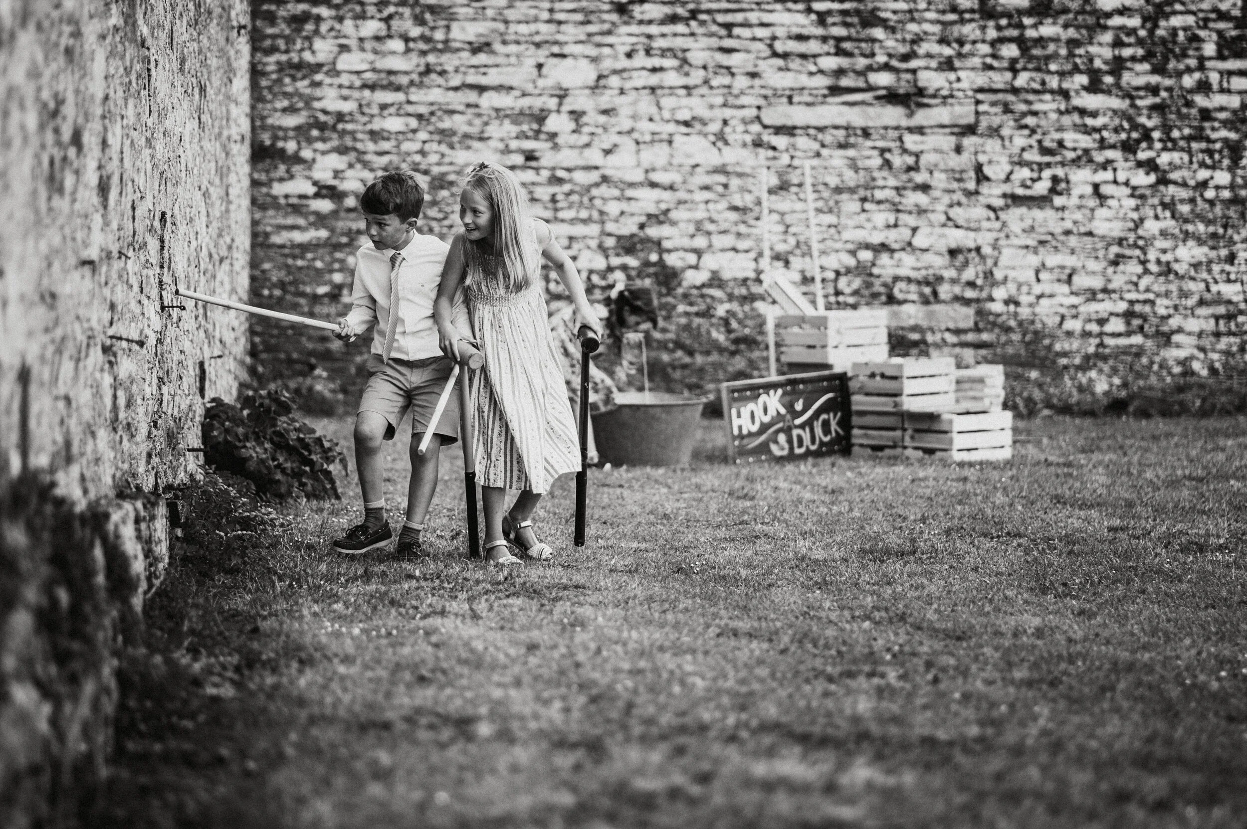 Two children, a boy and a girl, playing outside in a yard with a stone wall background. The girl is using crutches and the boy is holding a fishing pole.