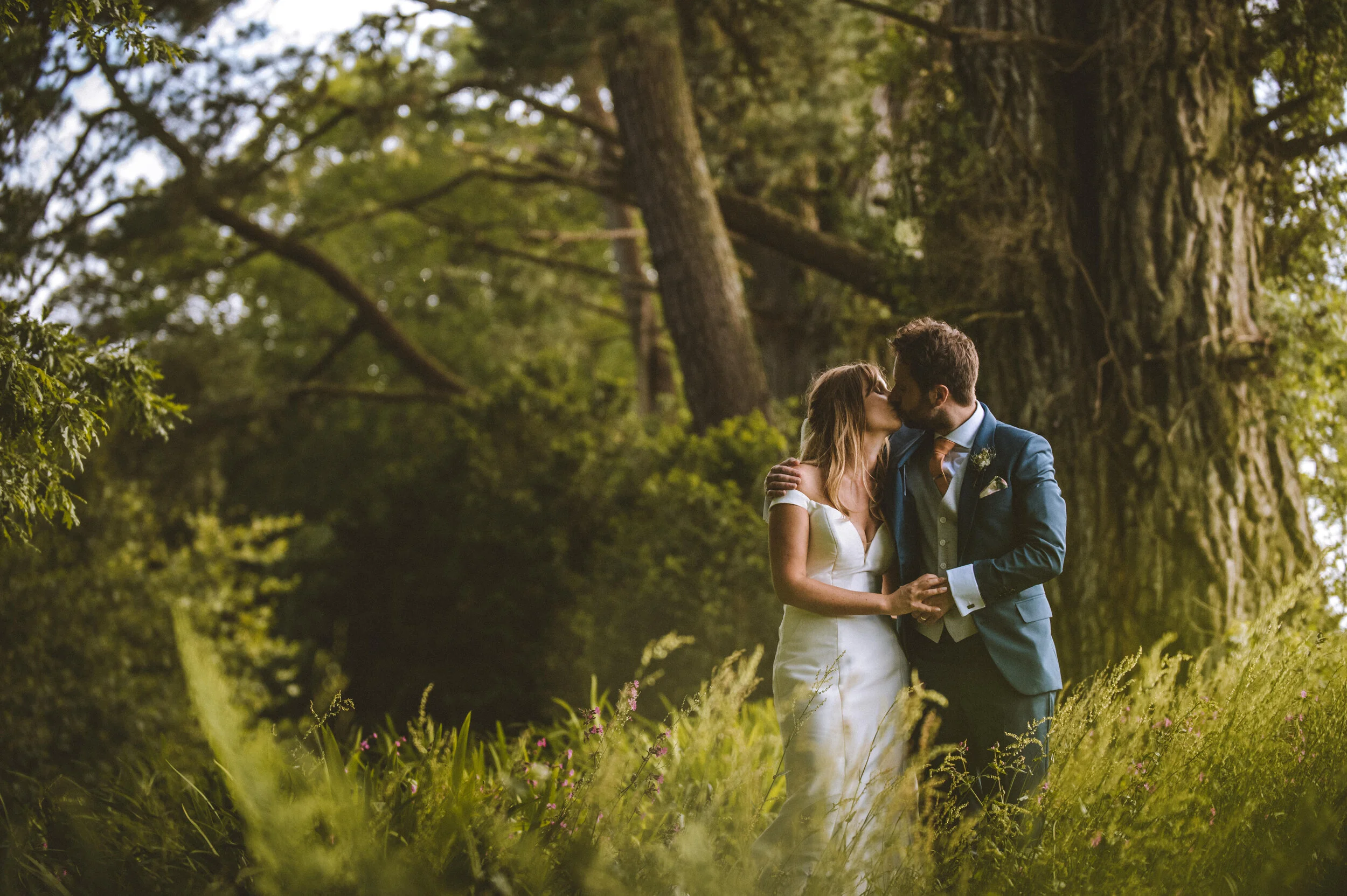 A couple in wedding attire kissing outdoors amid tall grass and trees.