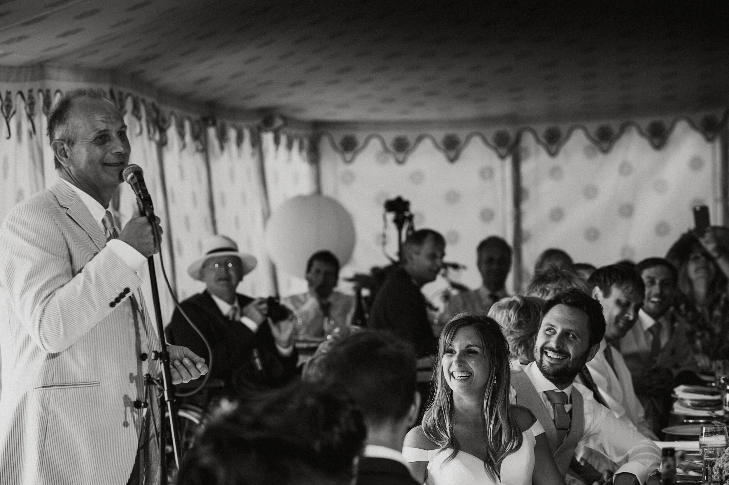 A man in a light-colored pinstripe suit giving a speech with a microphone at a wedding reception. Several guests are seated at the table, smiling and listening, some looking at the speaker and others looking at other guests. The scene is indoors with