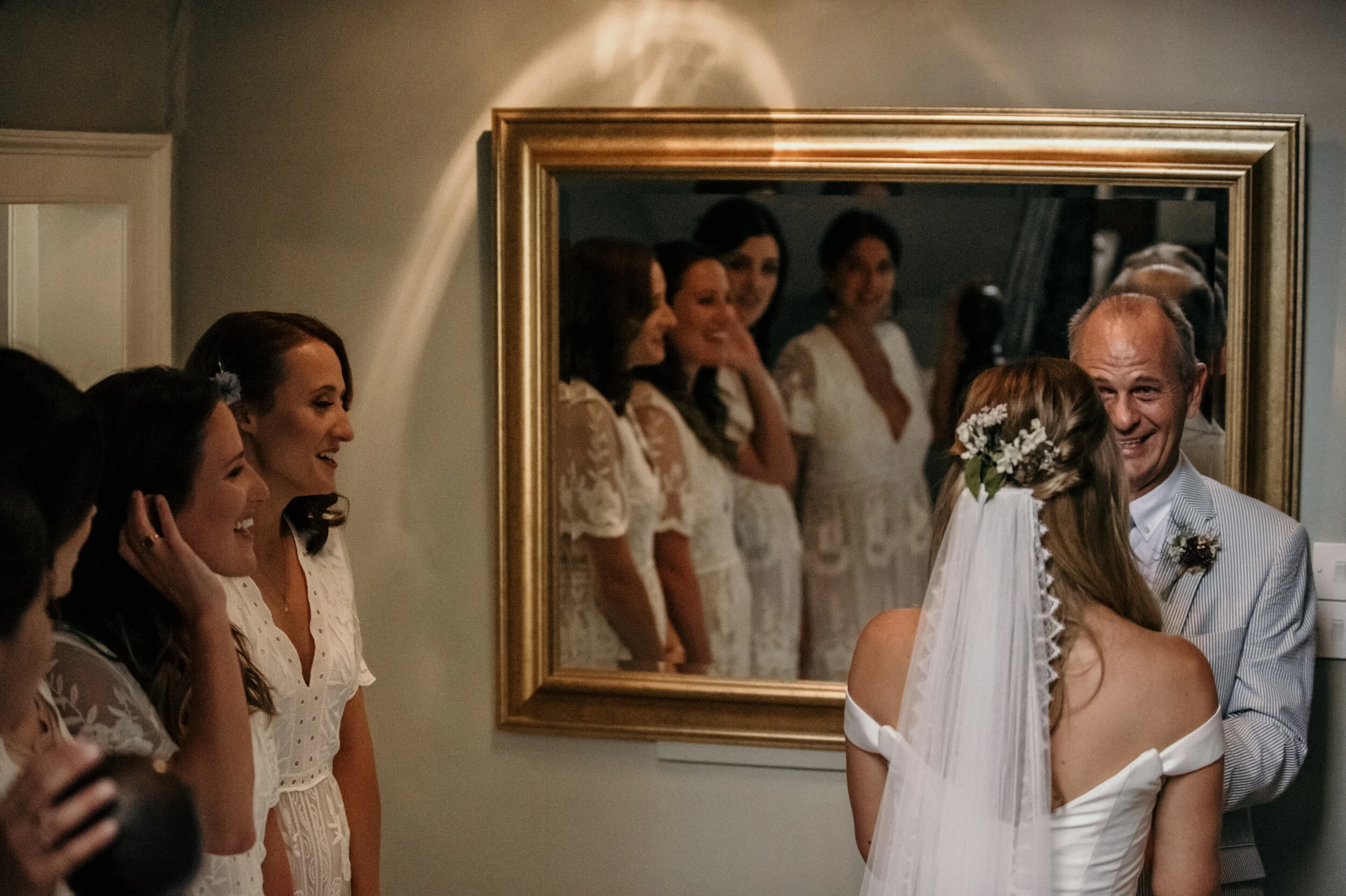 Bride and older man in a suit embracing in front of a mirror, with bridesmaids watching and smiling, in a warmly lit indoor setting.