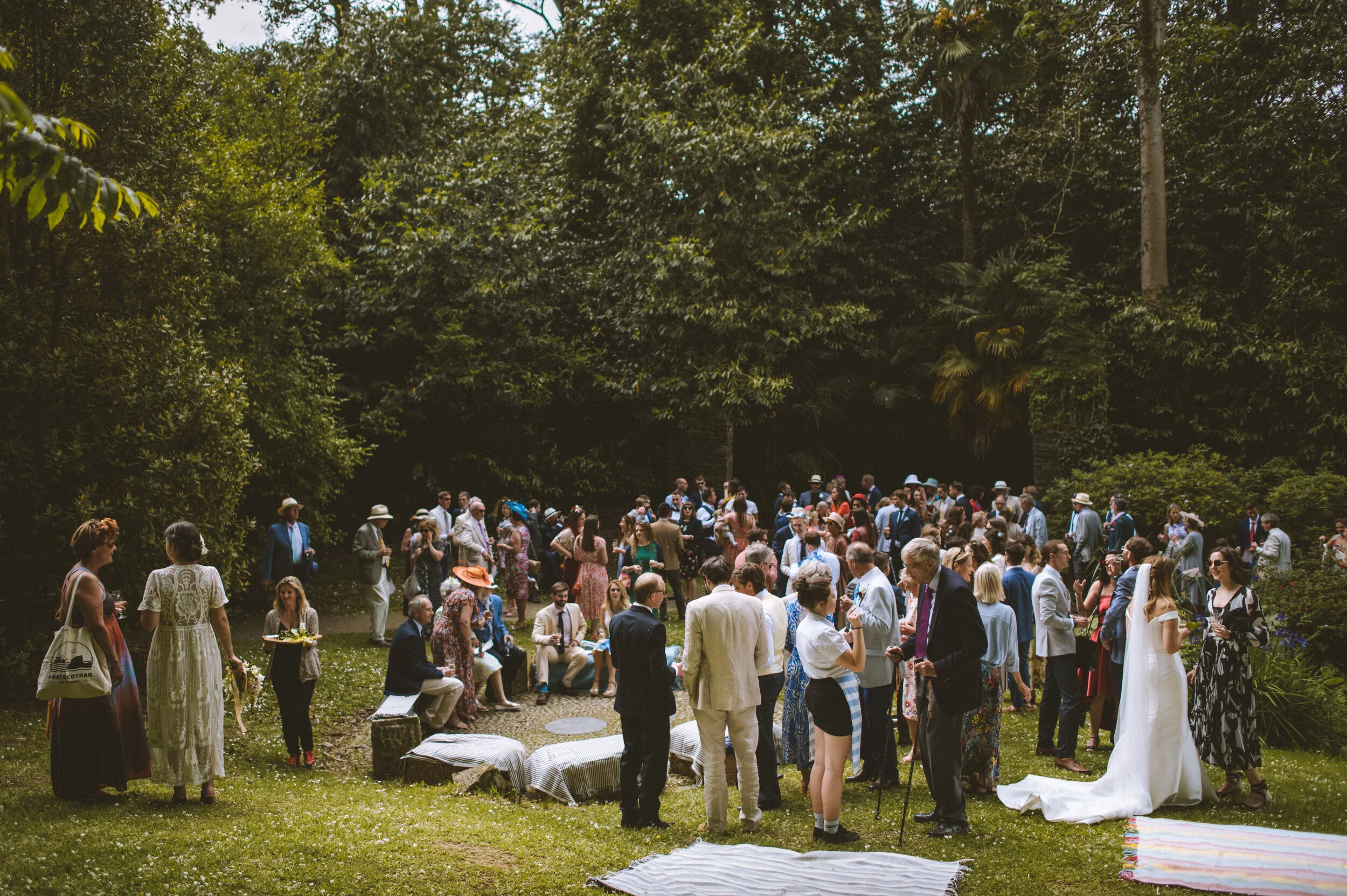 A large outdoor gathering of people dressed in formal attire, standing and sitting on a grassy area surrounded by trees, some wearing hats, with picnic blankets on the ground, likely a wedding or similar celebration.