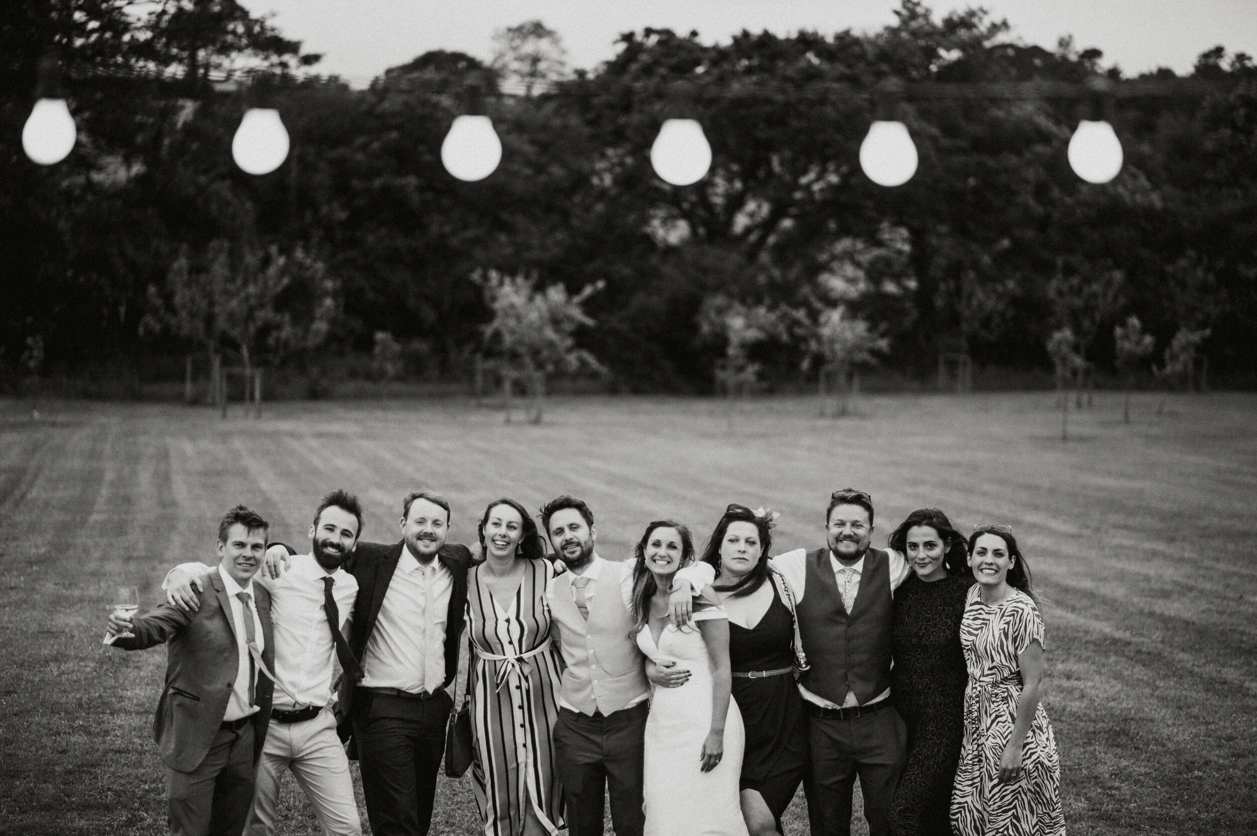 Black and white photo of a group of eleven people, dressed in formal and semi-formal attire, standing on a grassy field with trees in the background. They are smiling, with some having arms around each other, appearing to be celebrating an event or g