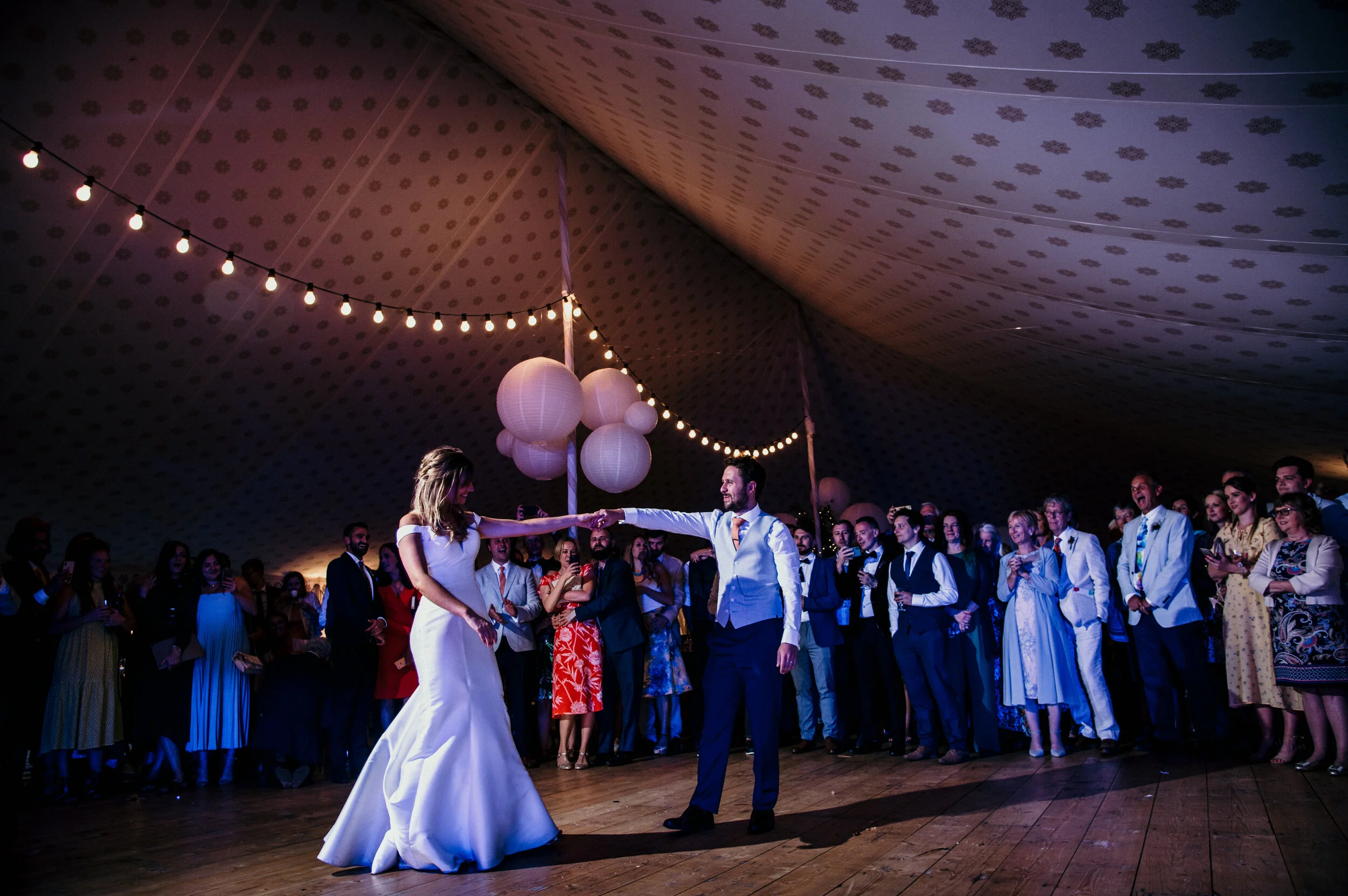 A bride and groom dancing at their wedding reception, surrounded by guests under a decorated tent with hanging lanterns and string lights.