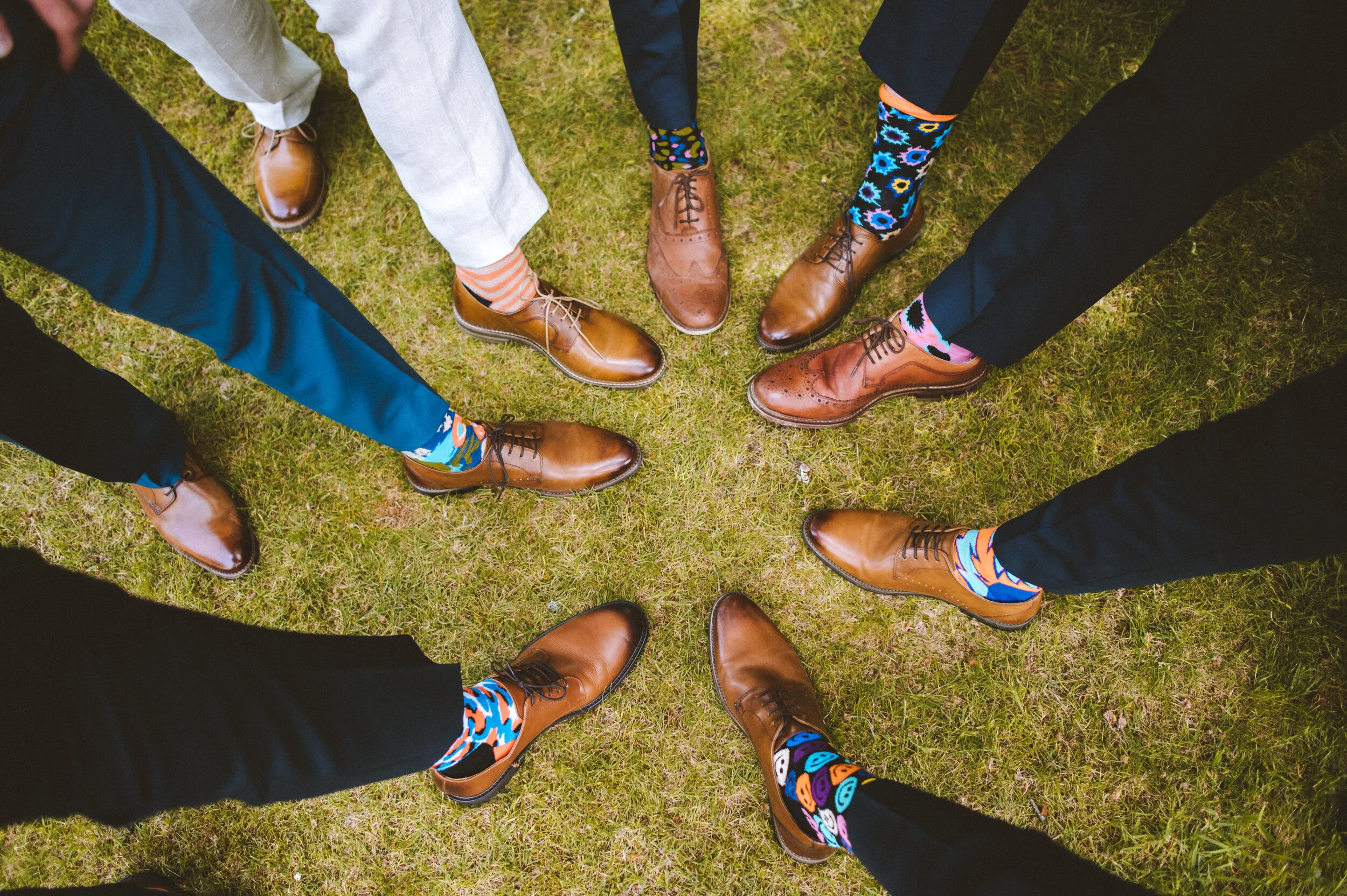 Multiple people standing in a circle on grass, wearing brown dress shoes and colorful patterned socks.