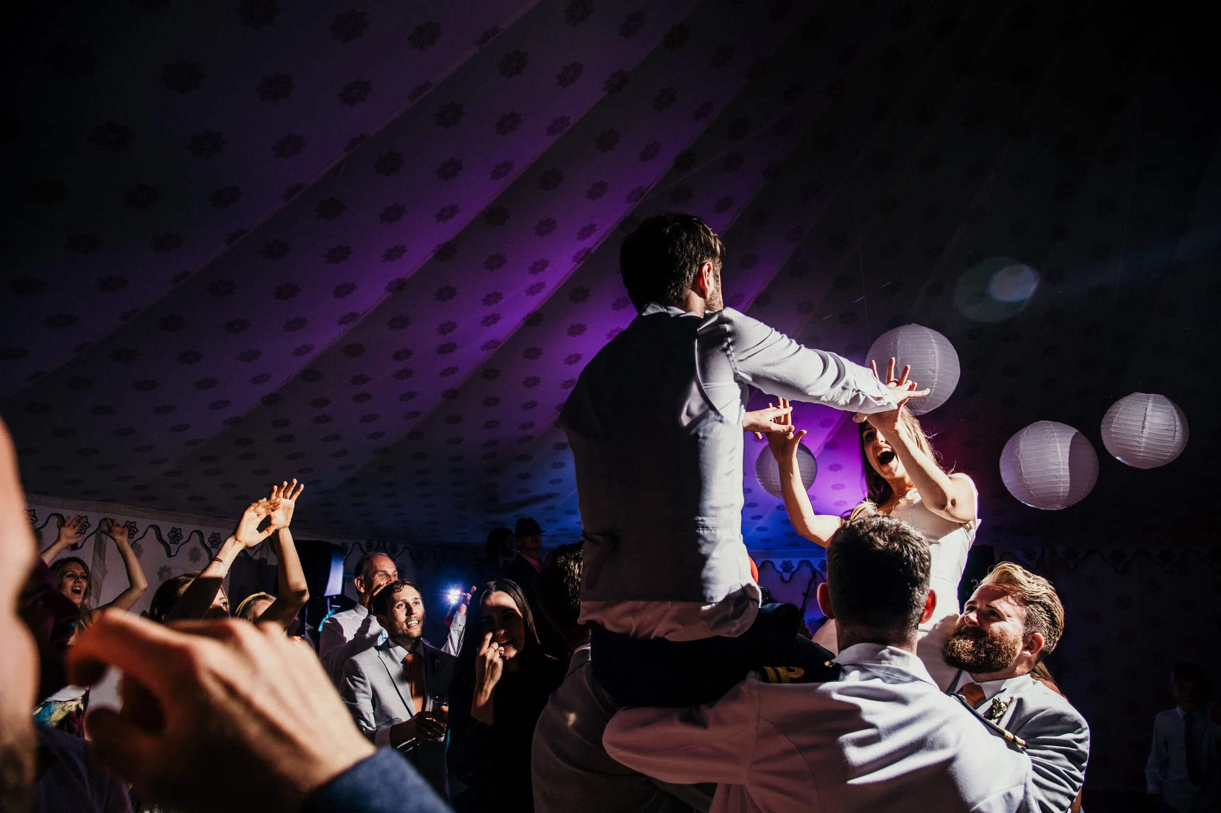People celebrating at a lively event, with one man being lifted on someone's shoulders and two women cheering in the background under illuminated paper lanterns.