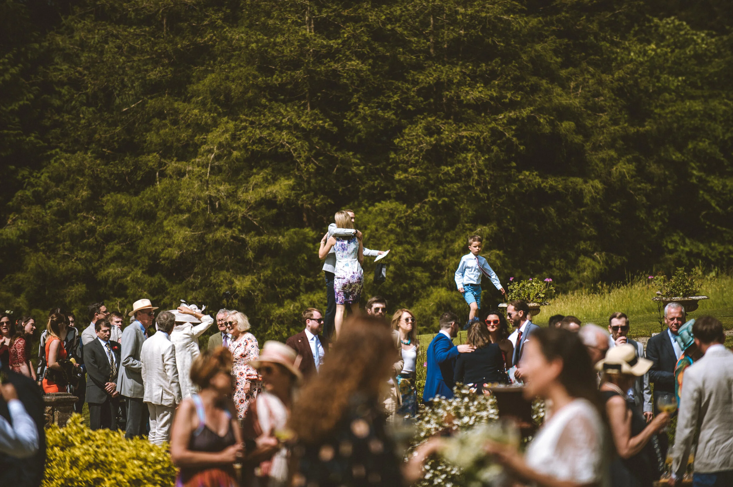 Pencarrow House outdoor social gathering with many people dressed in formal and casual attire, some wearing hats, set against a green, wooded background, with a young boy and a woman in the center, and flowers and plants surrounding the scene.